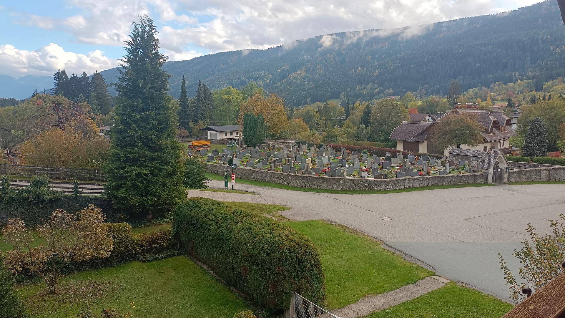Weitläufiger Blick auf eine ländliche Umgebung mit Friedhof und bewaldeten Bergen im Hintergrund.