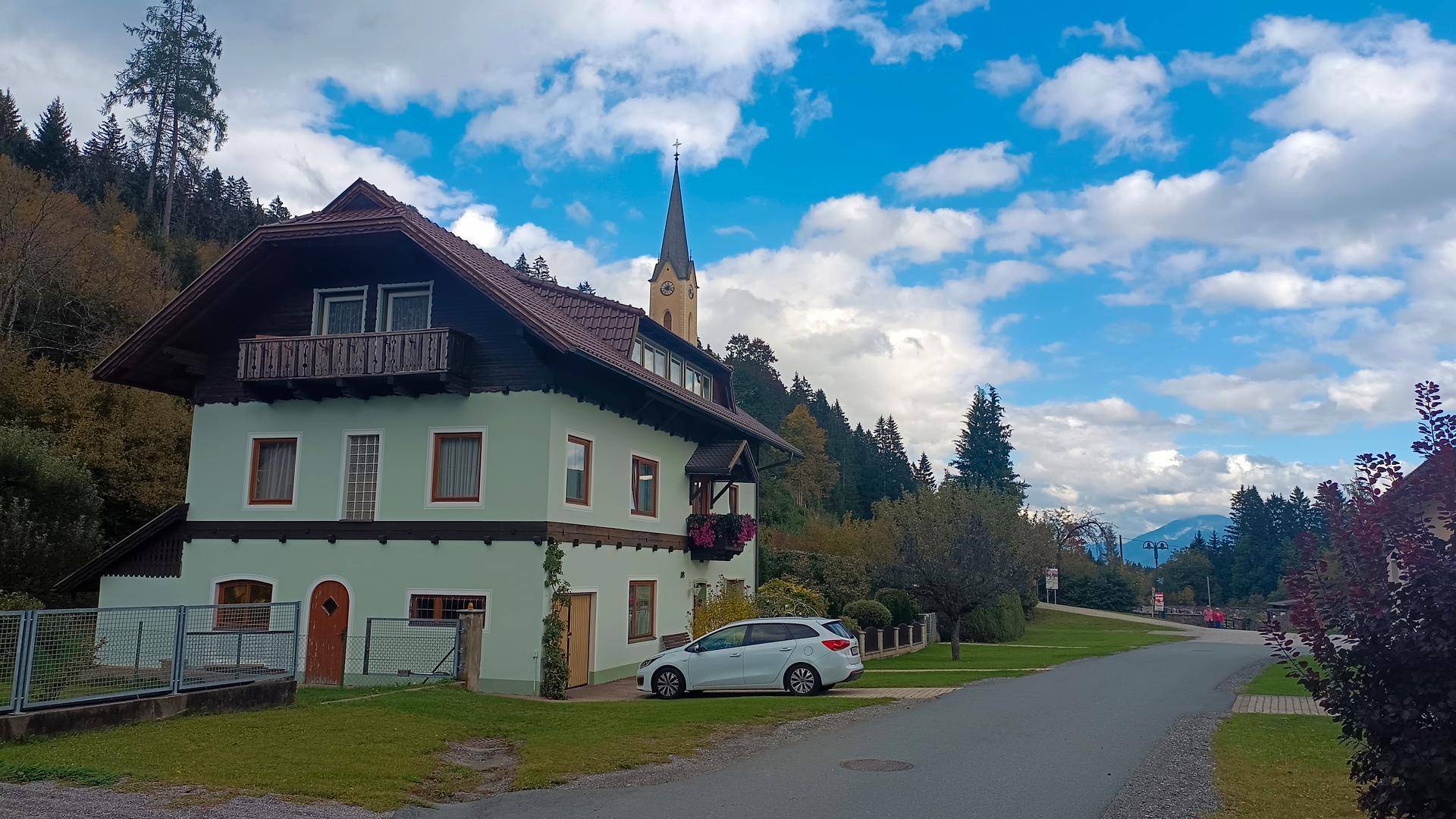 Haus in ländlicher Umgebung mit Kirchturm im Hintergrund und bewölktem Himmel.