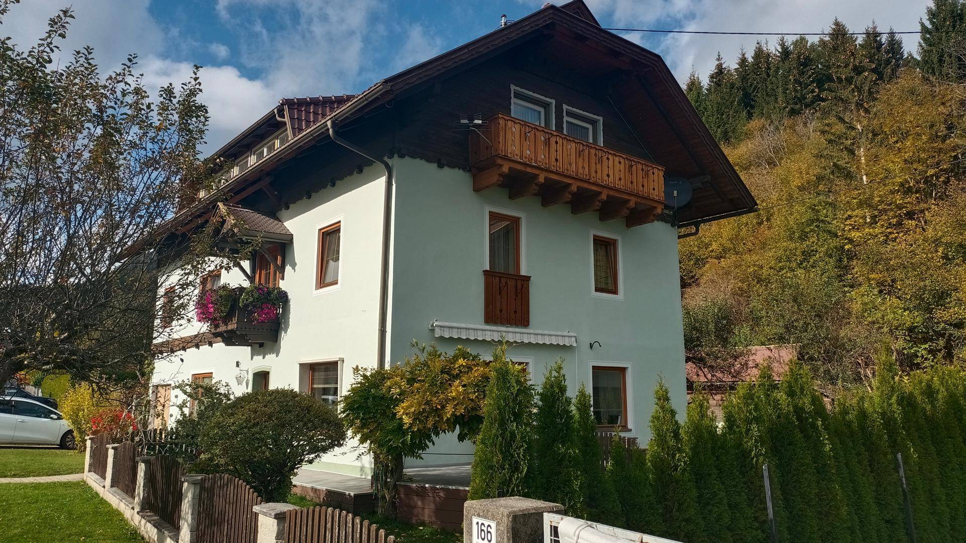 Ländliches Haus mit Holzbalkonen und grüner Hecke im Vordergrund, umgeben von Bäumen.