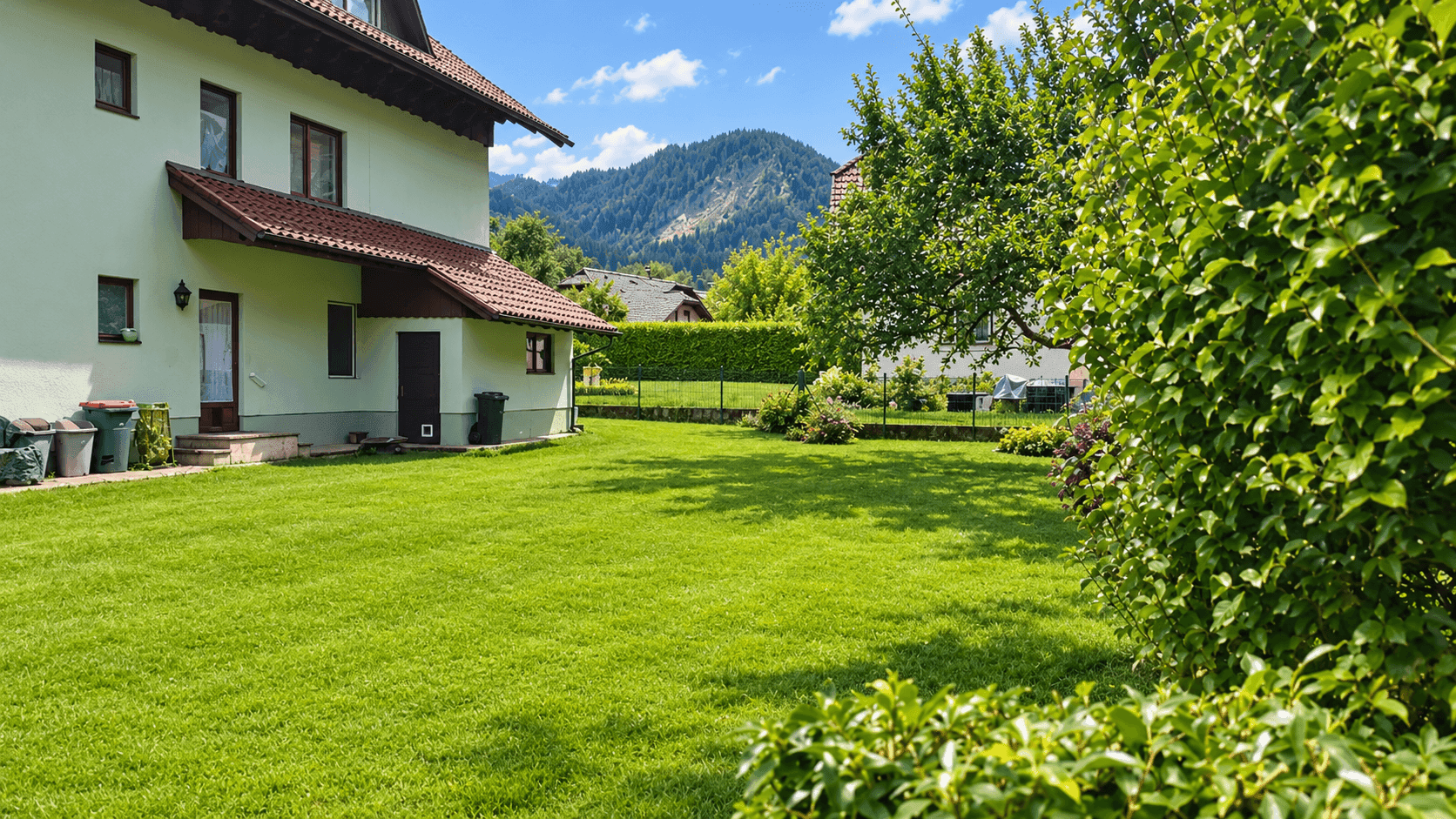 Weitläufiger, sonniger Garten mit Rasenfläche und Bäumen, umgeben von einer Berglandschaft.