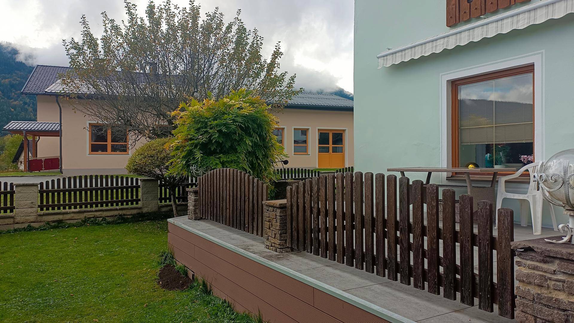 Grüner Gartenbereich mit Holzzaun und Blick auf ein Nachbargebäude und die umliegende Landschaft.