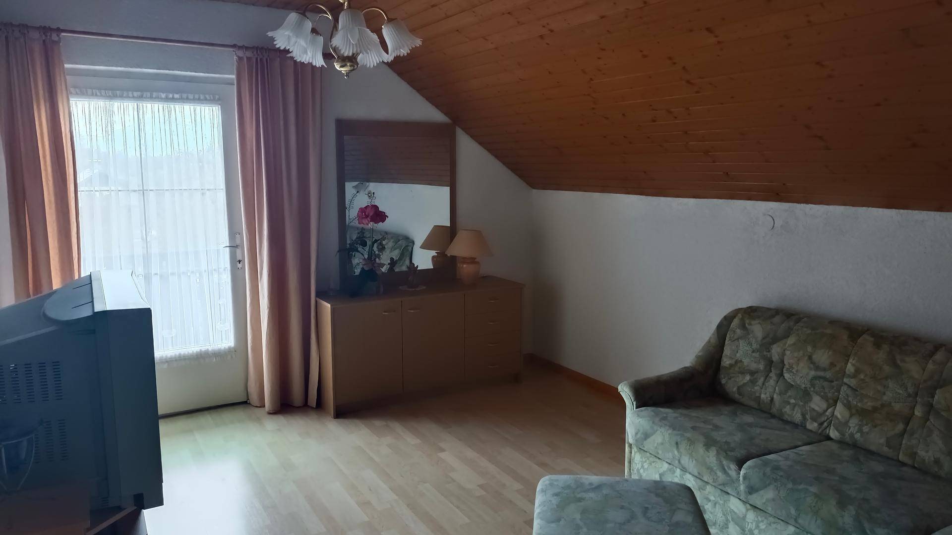 Gemütliches Wohnzimmer im Dachgeschoss mit Holzdecke, Sofa und Zugang zum Balkon.