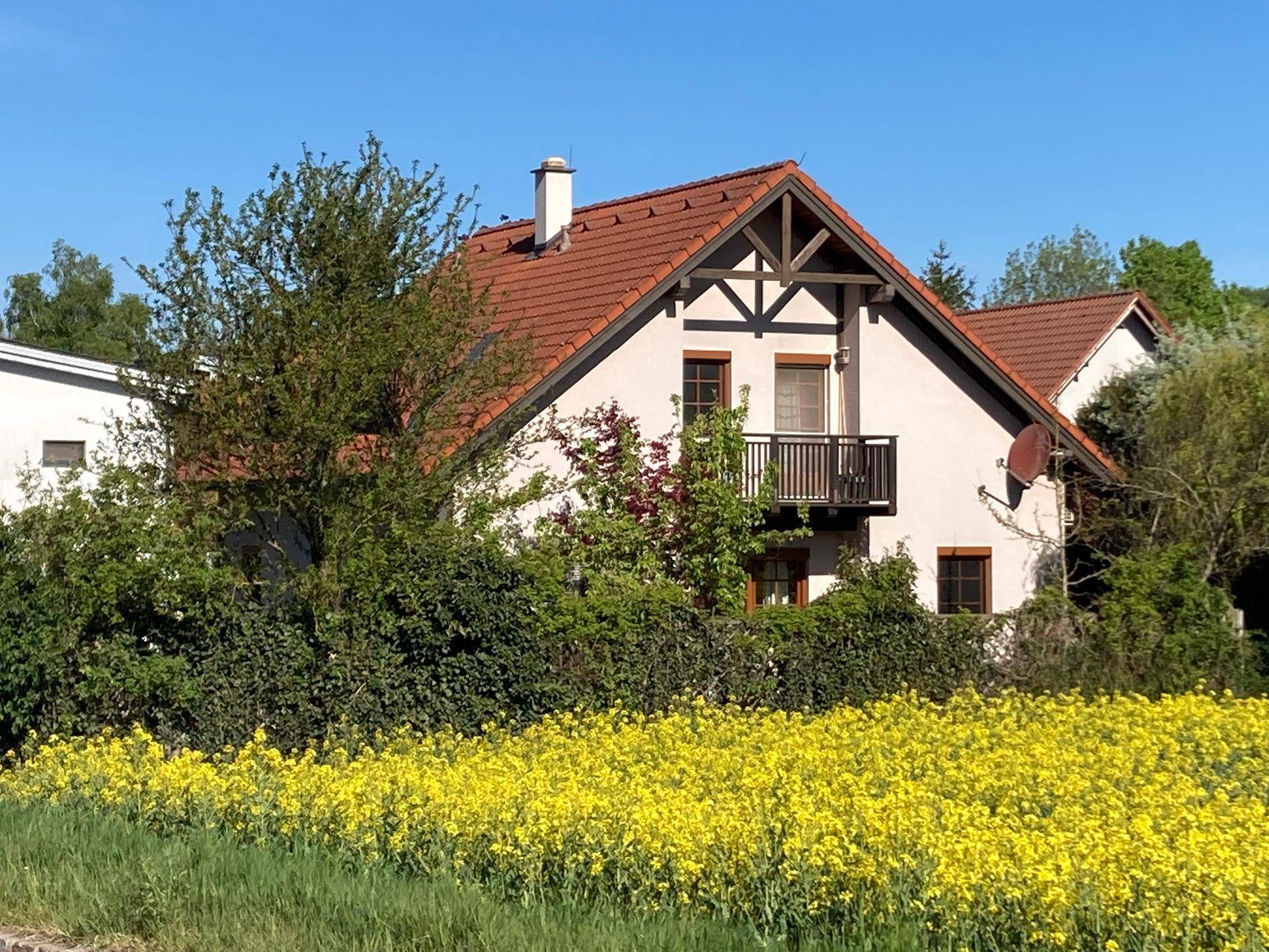 Hausfassade mit kleinem Balkon, umgeben von üppigem Grün und einem blühenden Rapsfeld im Vordergrund.