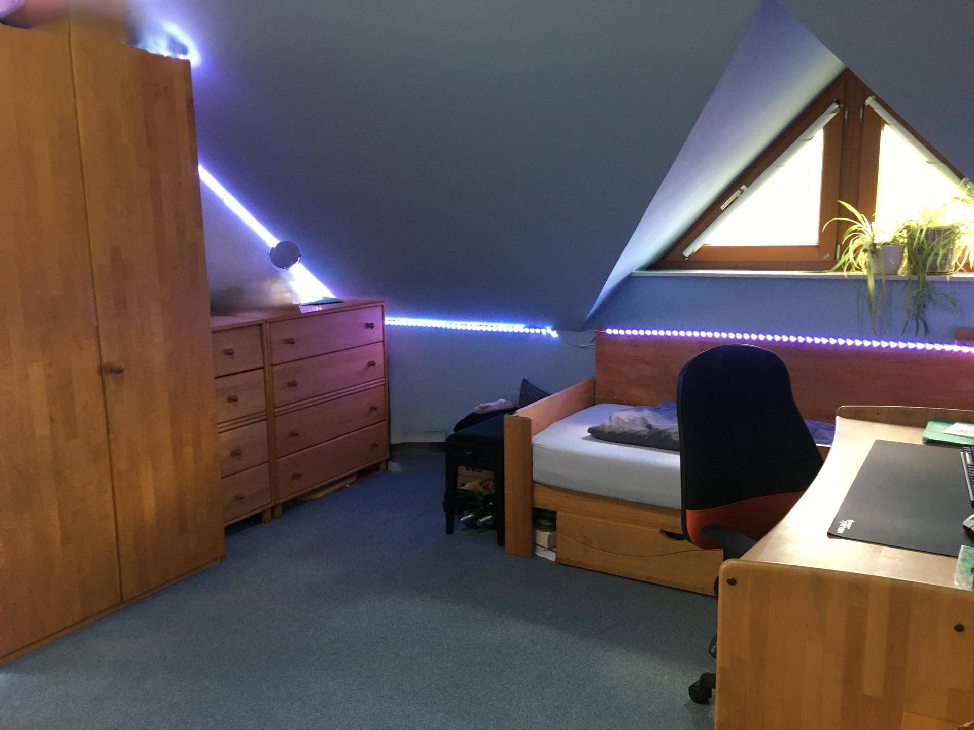 Schlafzimmer mit Dachschräge, dreieckigem Fenster, Holzmöbeln und blauer Teppichboden.