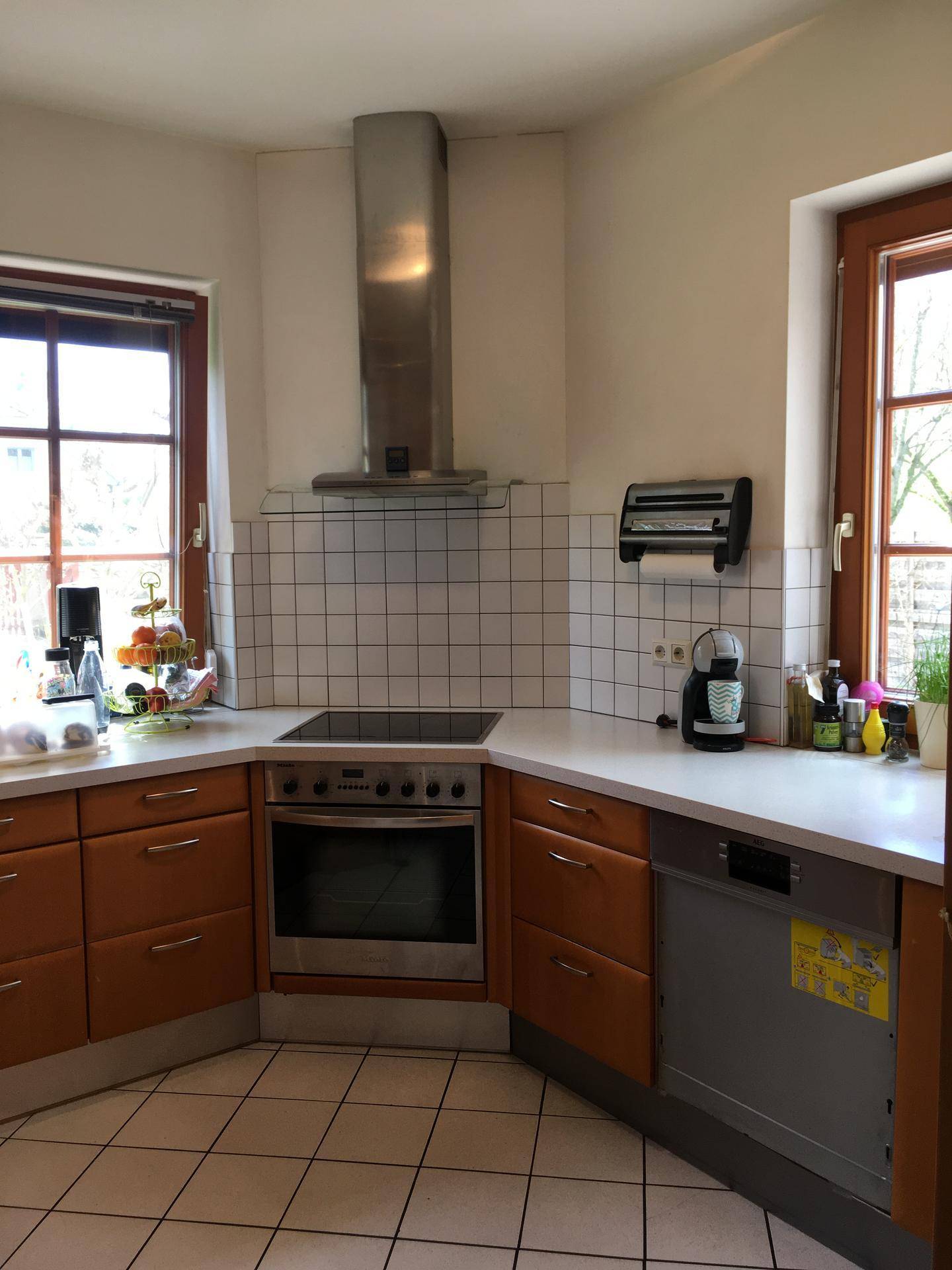 Funktionale Küche mit Holzschränken, weißen Arbeitsplatten, Fliesenboden und zwei Fenstern.