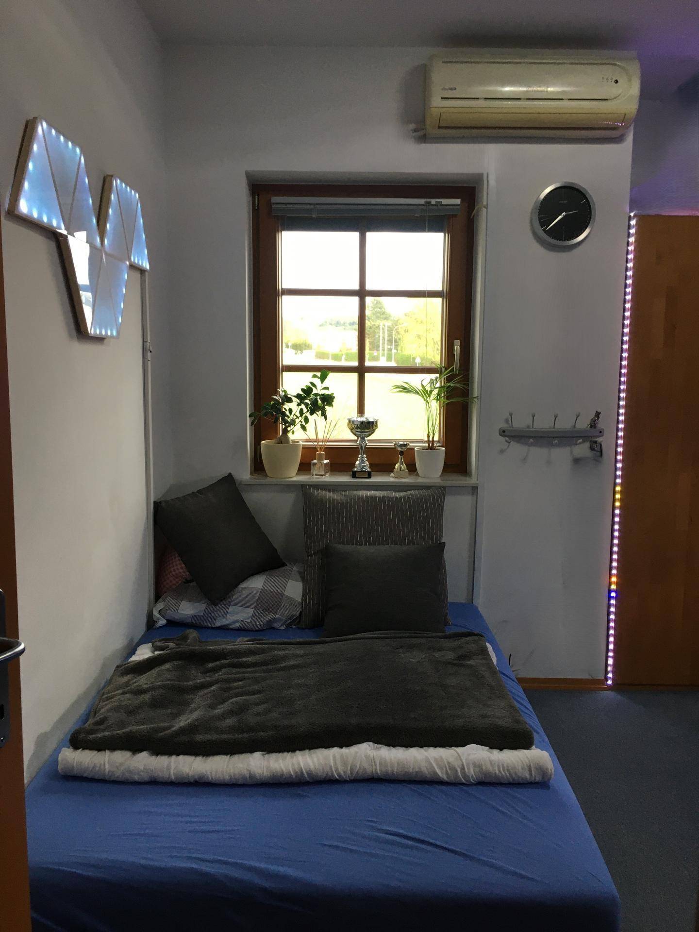 Helles Schlafzimmer mit Einzelbett, Fenster, Klimaanlage und blauer Teppichboden.