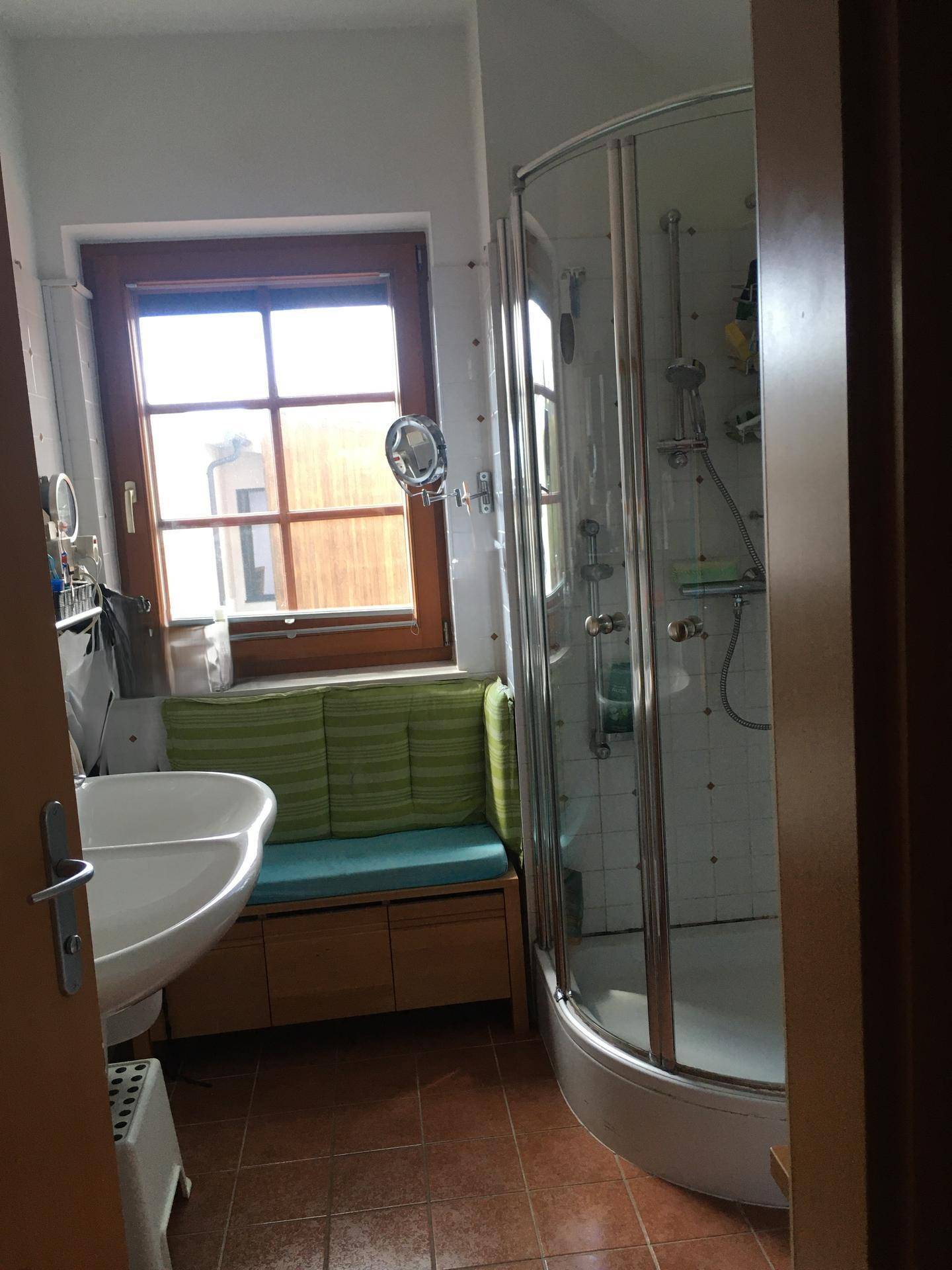 Badezimmer mit Eckdusche, Waschbecken, Fenster und terracottafarbenen Fliesenboden.