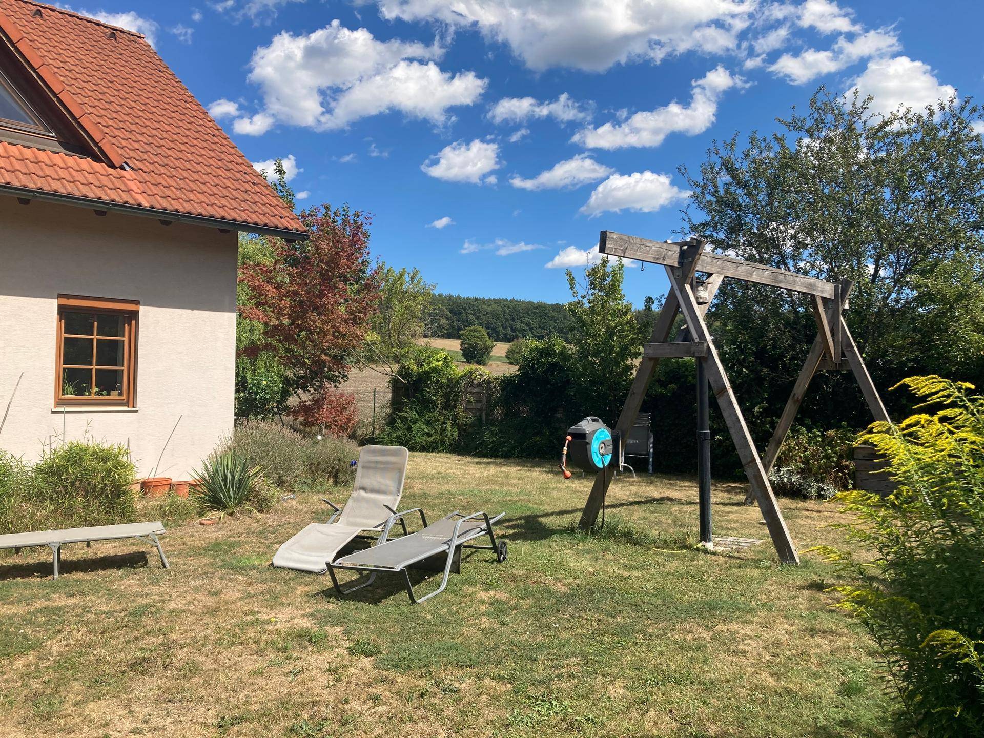 Sonniger Gartenbereich mit Liegestühlen und einer Schaukel, umgeben von Natur und Hügellandschaft.