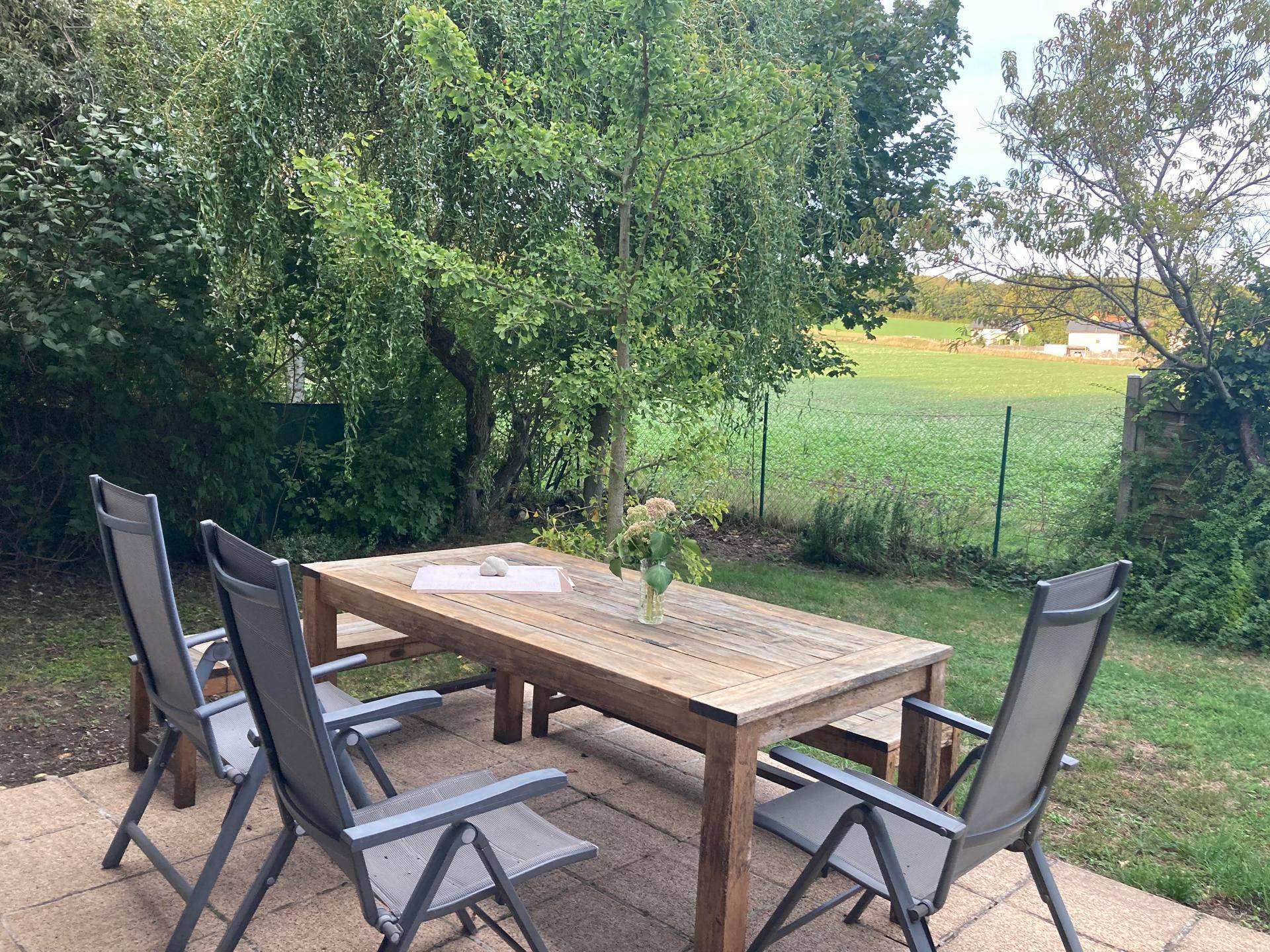 Gemütlicher Außenbereich mit Holztisch und Stühlen auf einer gepflasterten Terrasse, umgeben von Bäumen.