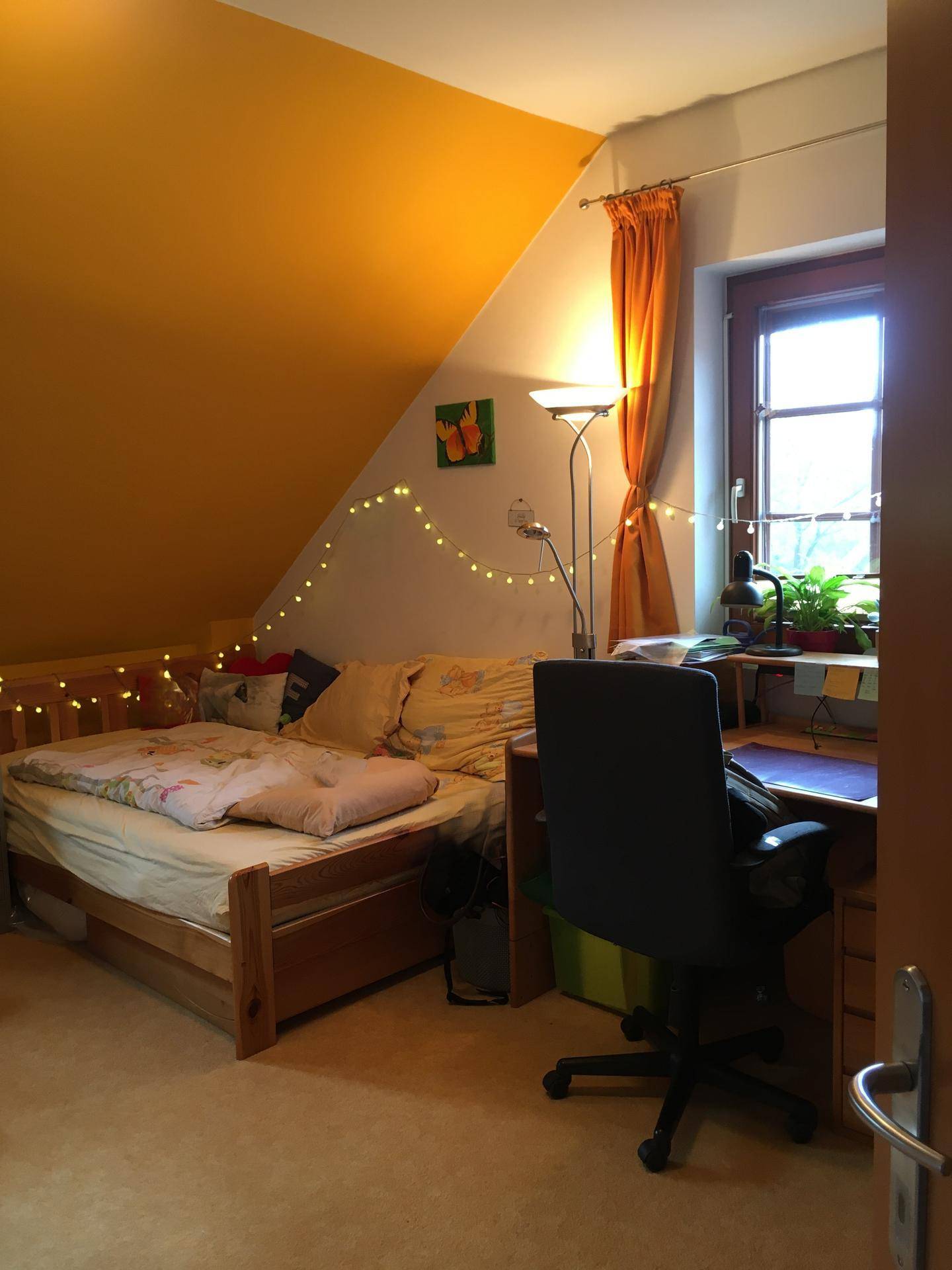 Schlafzimmer mit Einzelbett, Schreibtisch, Fenster und gelben Wänden, dekoriert mit Lichterketten.