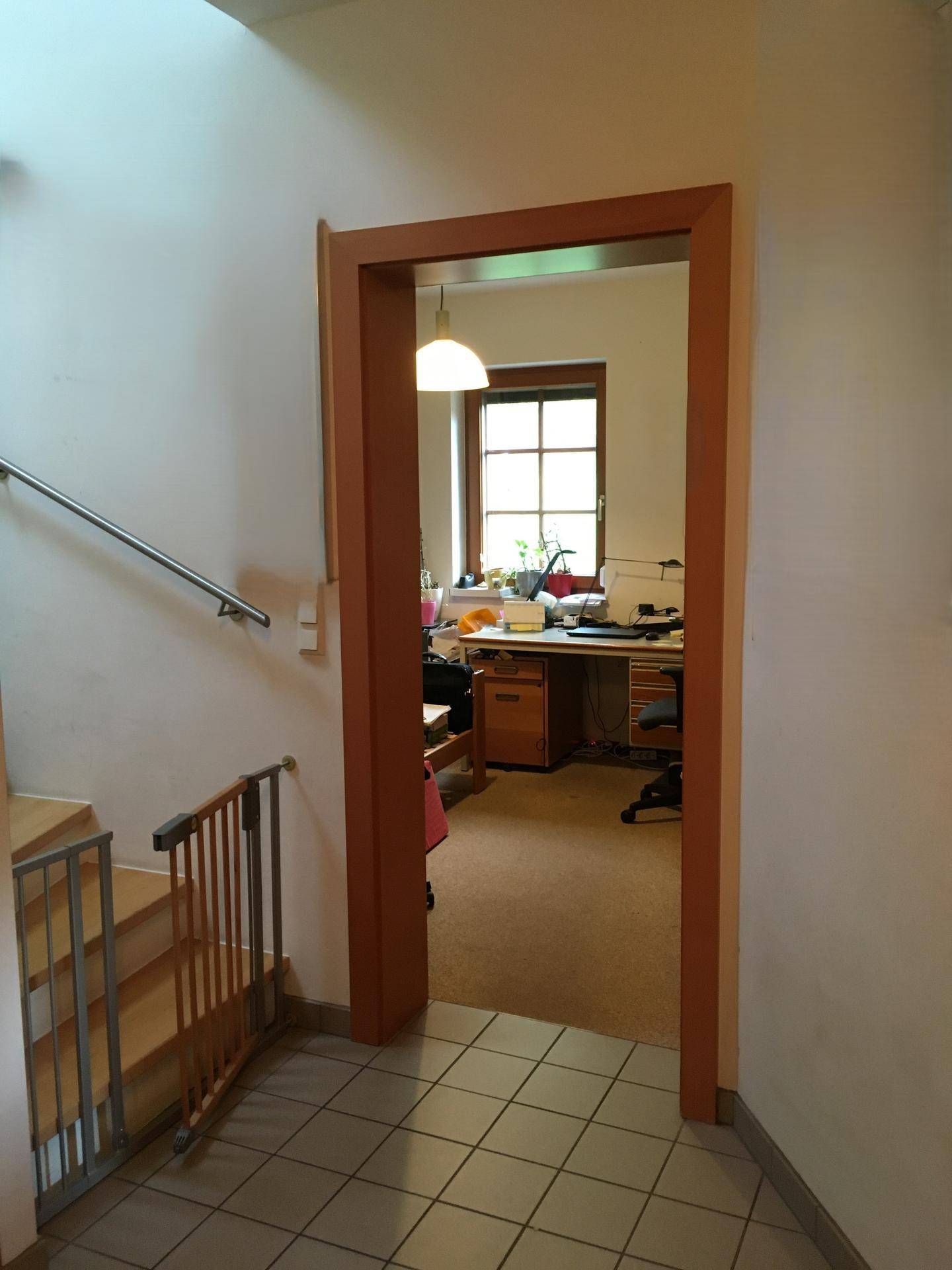 Heller Flur mit Fliesenboden, Treppe und Blick in ein angrenzendes Arbeitszimmer mit Fenster.