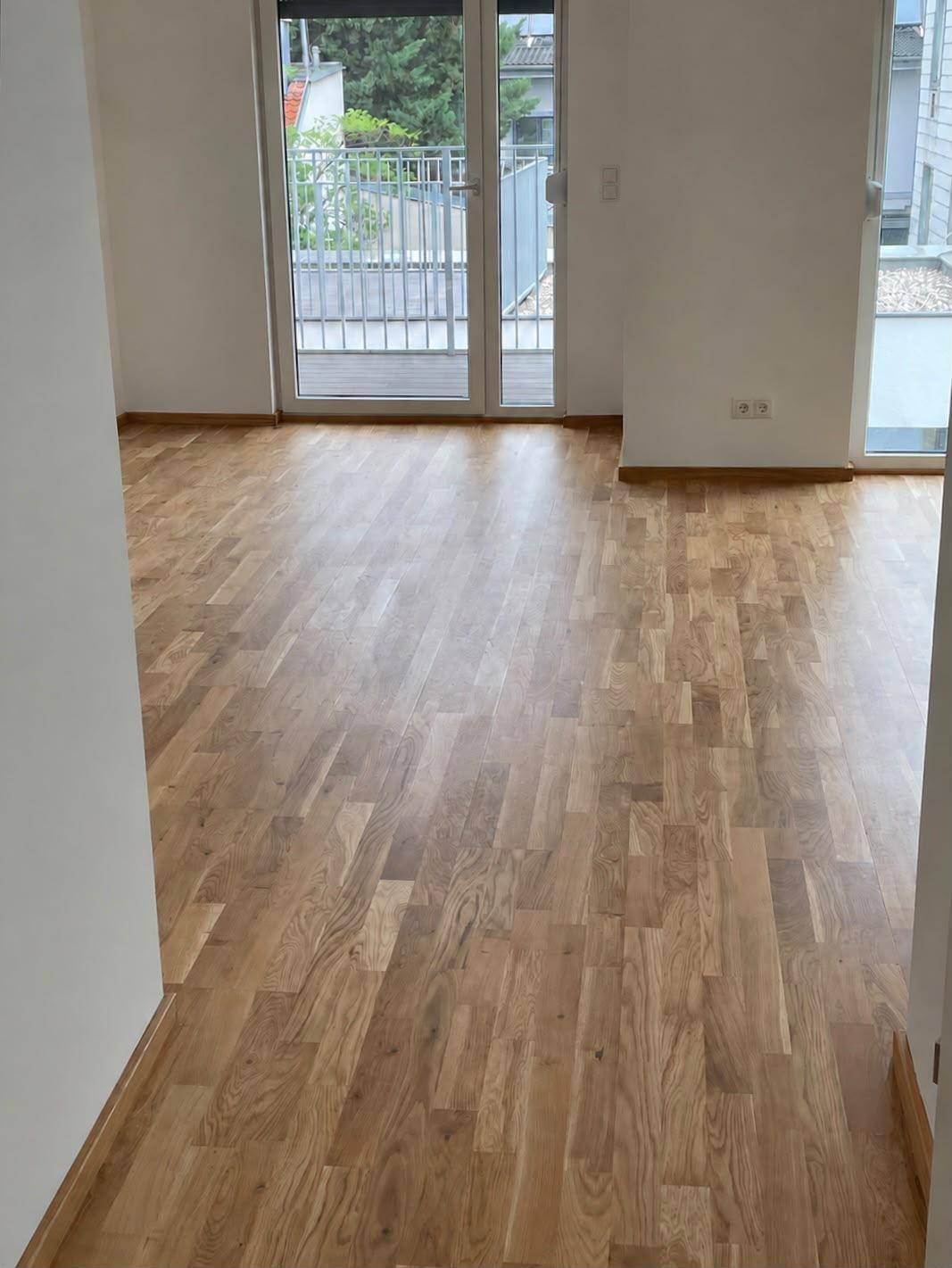 Leerer Raum mit hellem Holzboden und breiter Glastür, die zu einem Balkon oder einer Terrasse führt.