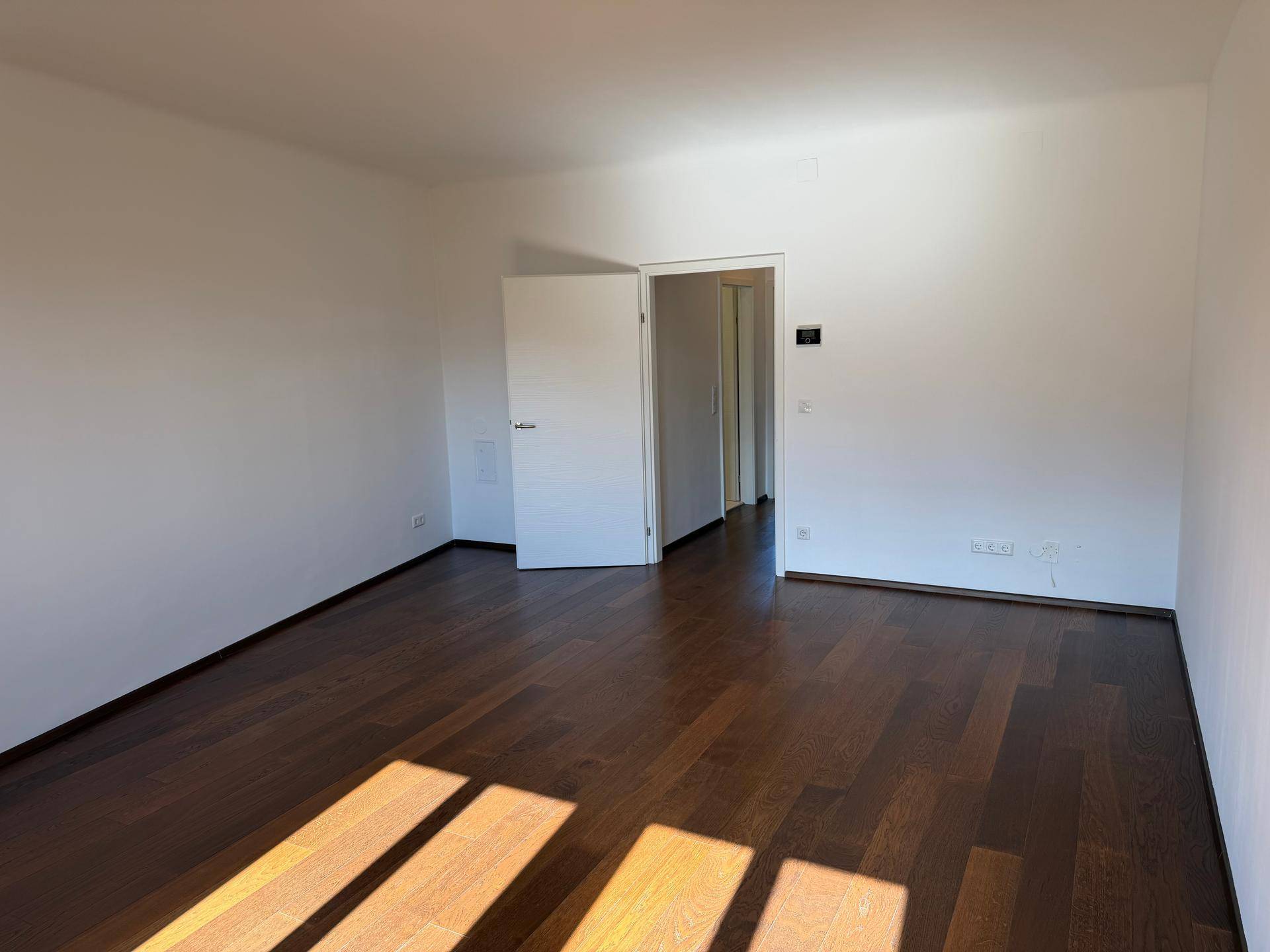 Geräumiges Zimmer mit dunklem Parkettboden und offener Tür zu einem weiteren Raum.