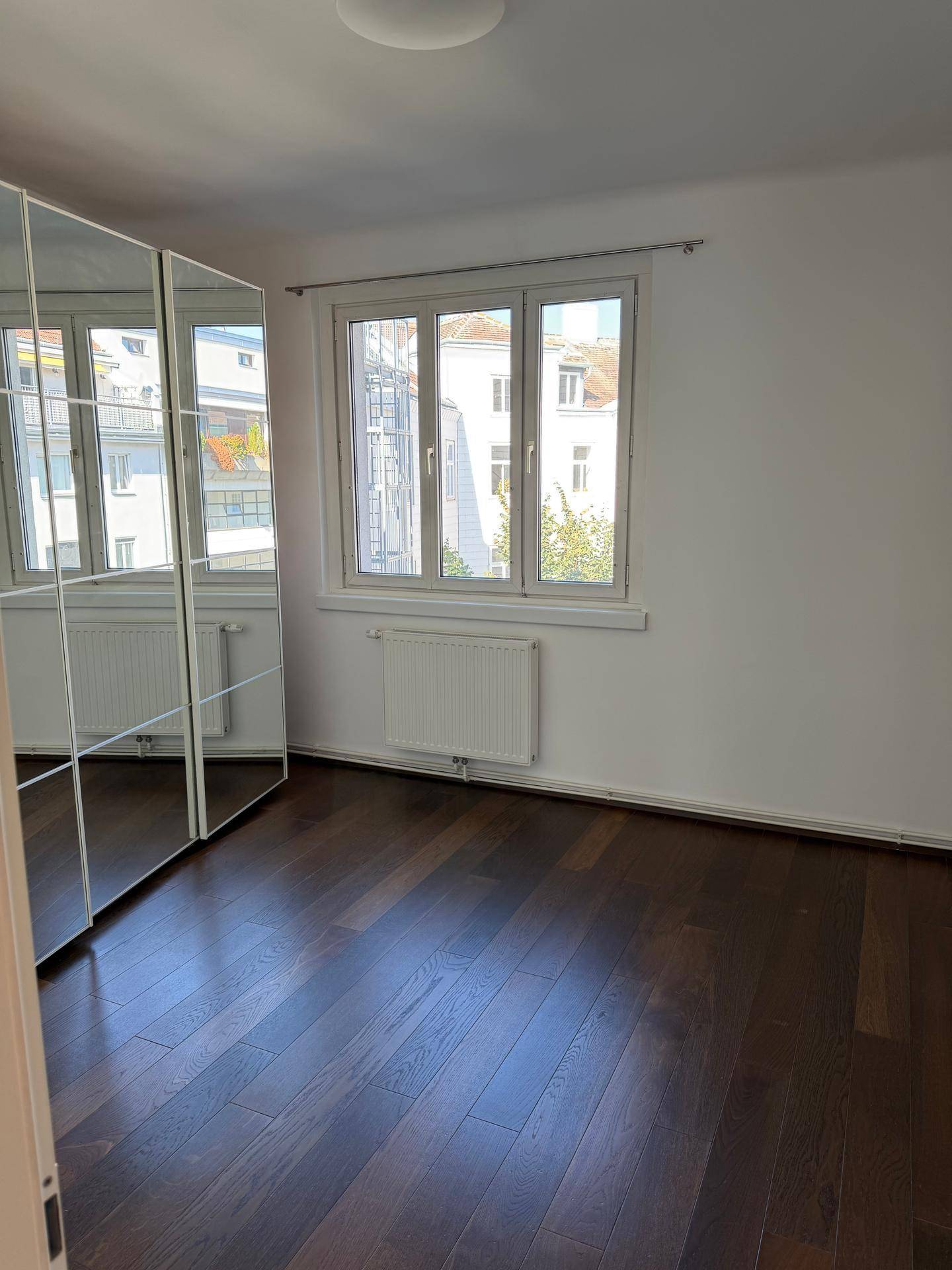 Helles Schlafzimmer mit dunklem Parkettboden, Fenster und einem großen Spiegelschrank.