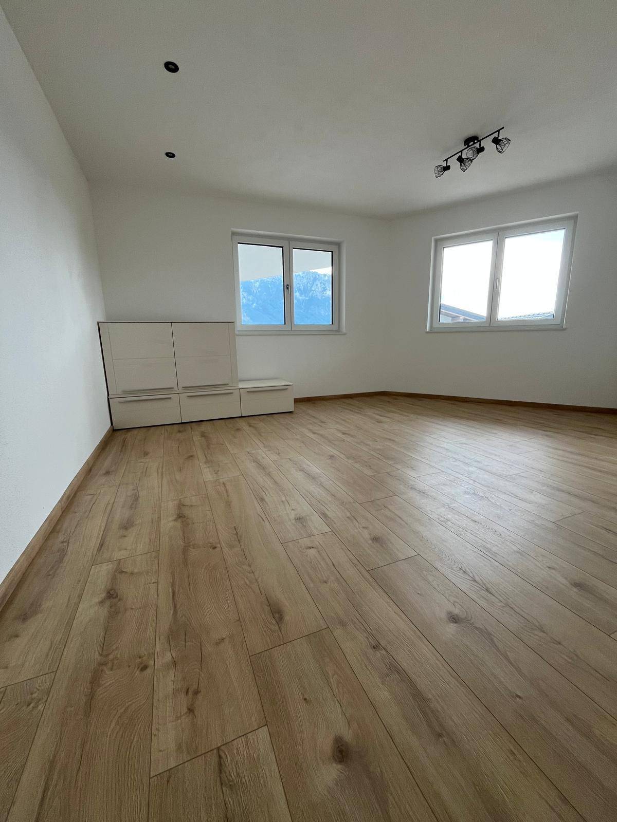 Geräumiger, heller Raum mit Laminatboden, großen Fenstern und einem niedrigen Schrank.