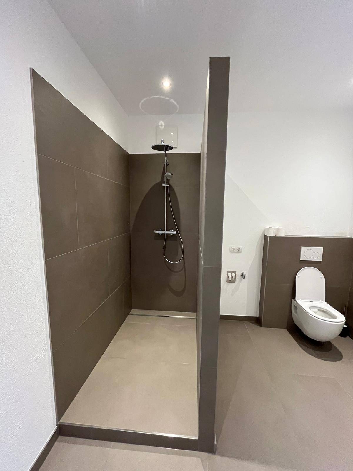 Modernes Badezimmer mit ebenerdiger Dusche, Wand-WC und großformatigen Fliesen in Erdtönen.