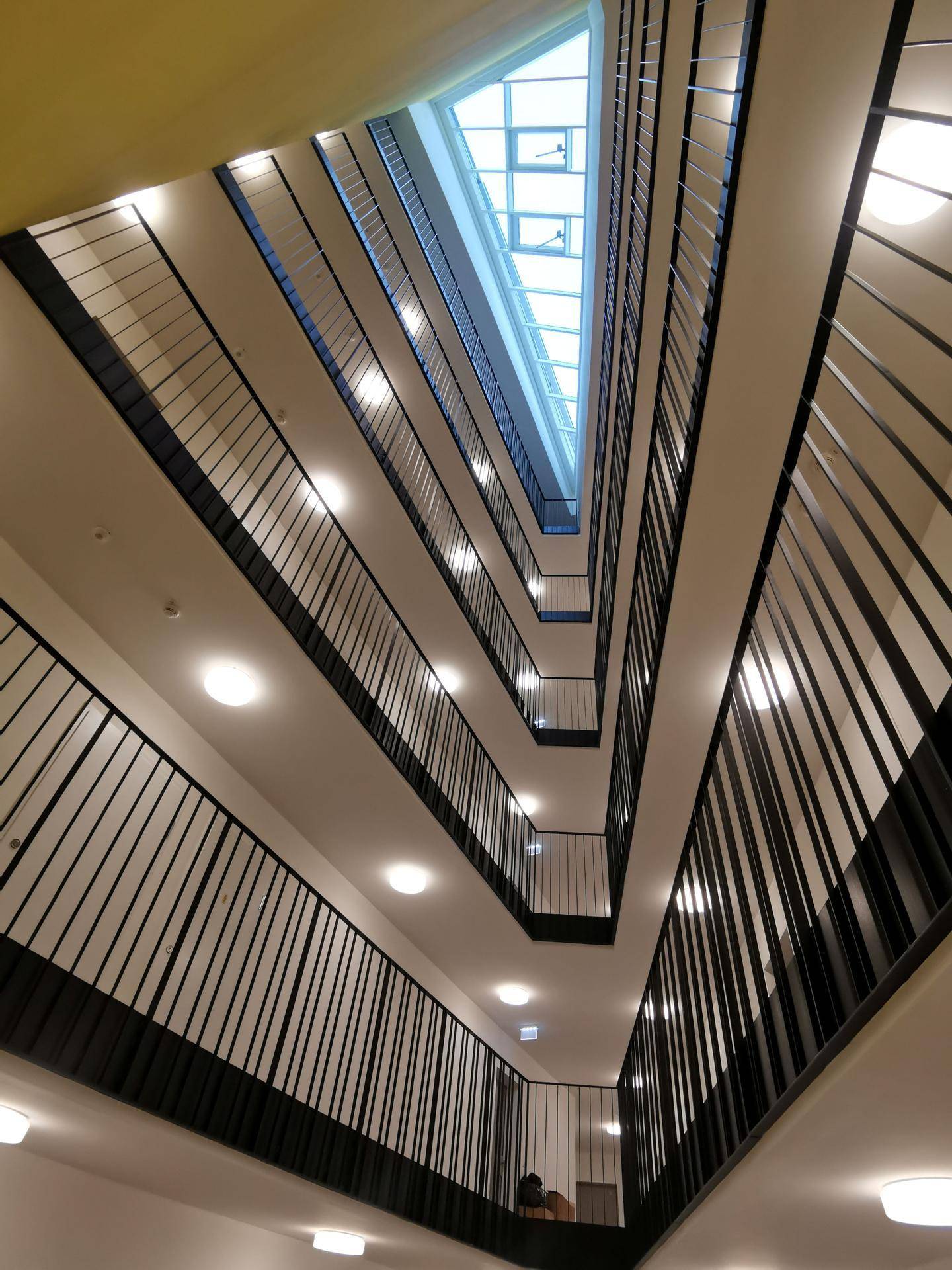 Blick nach oben in ein mehrstöckiges Treppenhaus mit Geländern und einem Oberlicht, das Tageslicht spendet.