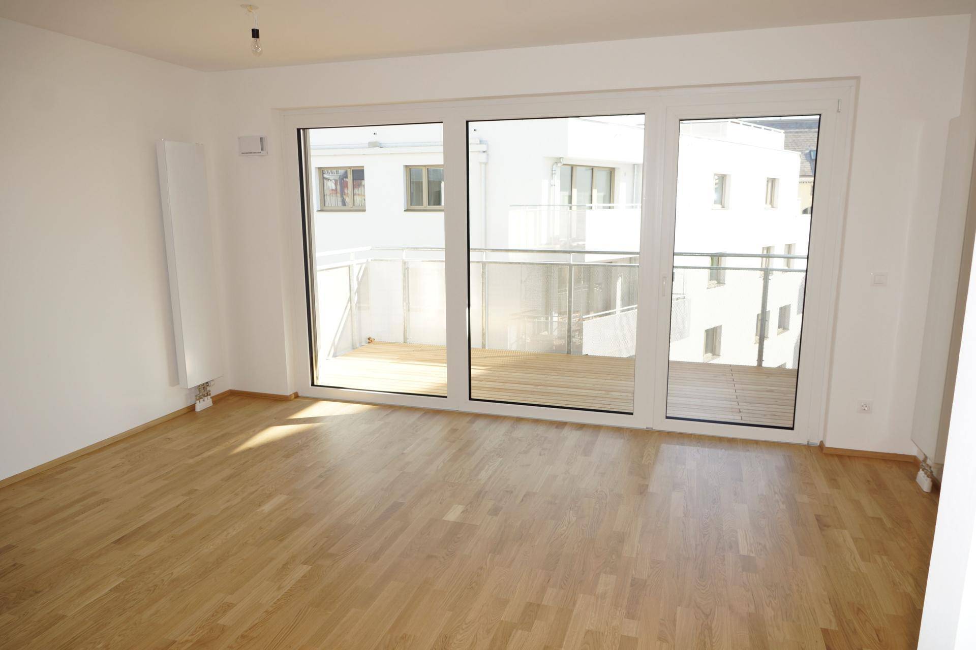 Geräumiges, unmöbliertes Zimmer mit hellem Holzboden und Zugang zu einem Balkon.