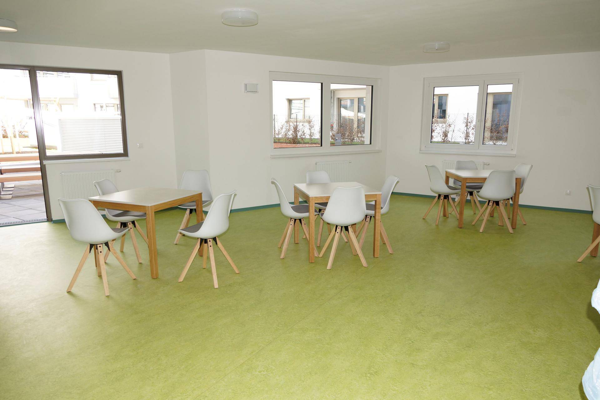 Heller Gemeinschaftsraum mit grünem Boden, mehreren Tischen und Stühlen, ideal für Treffen.