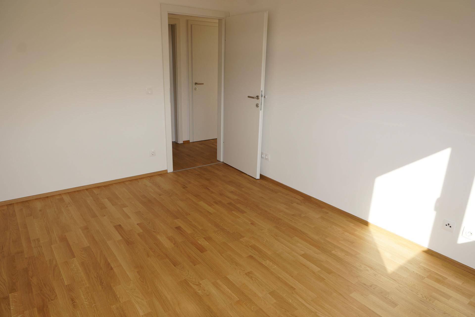 Leeres Zimmer mit Holzboden und weiß gestrichenen Wänden, durchflutet von natürlichem Sonnenlicht.