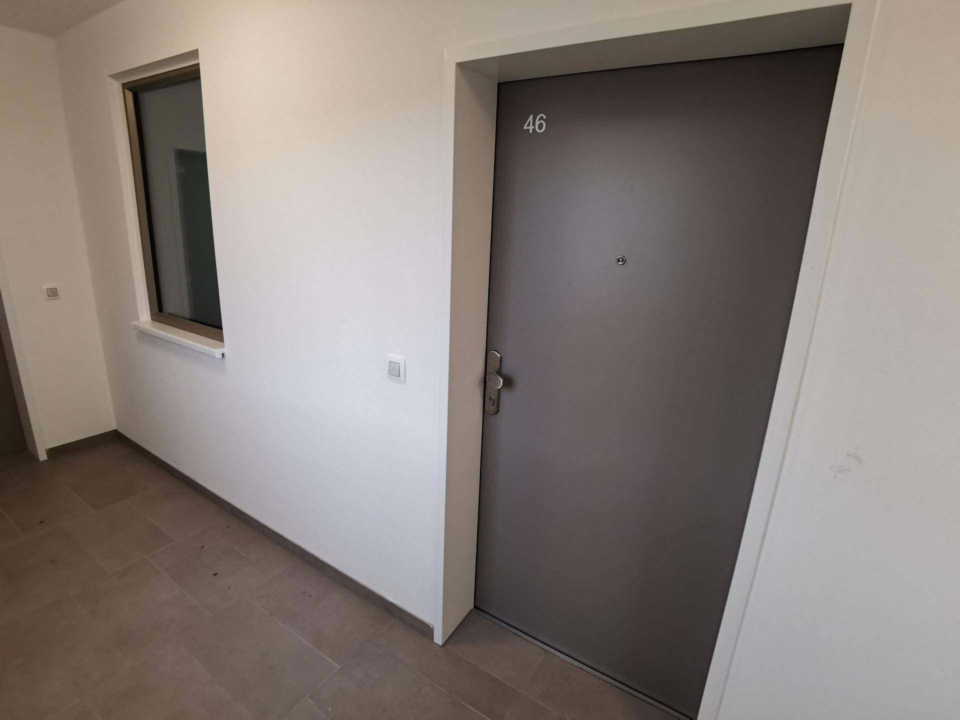 Heller Flur mit grauer Wohnungstür Nummer 46 und einem kleinen Fenster, ausgestattet mit Fliesenboden.
