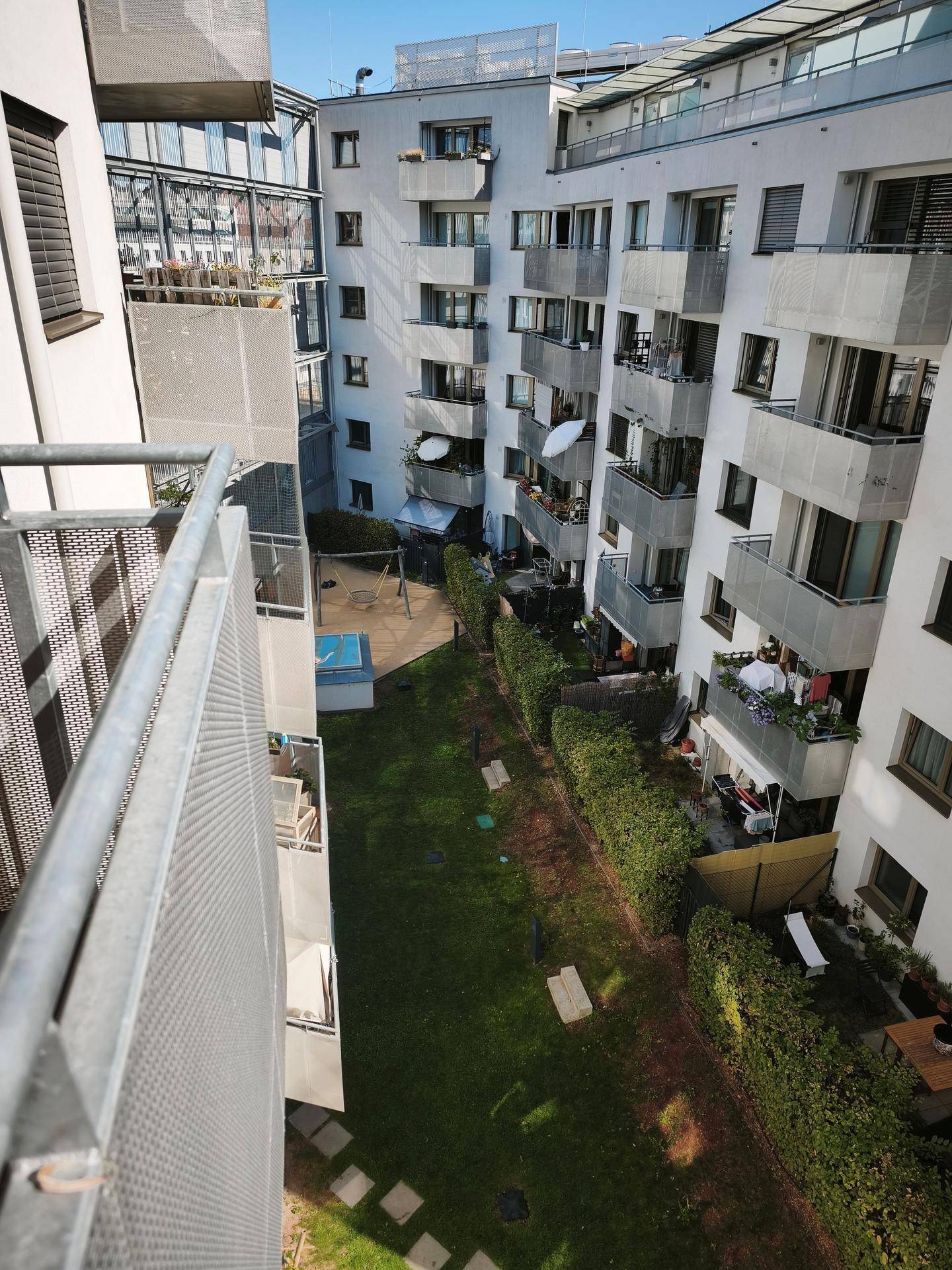 Blick von einem Balkon in einen Innenhof mit Rasenfläche, Hecken und weiteren Balkonen.