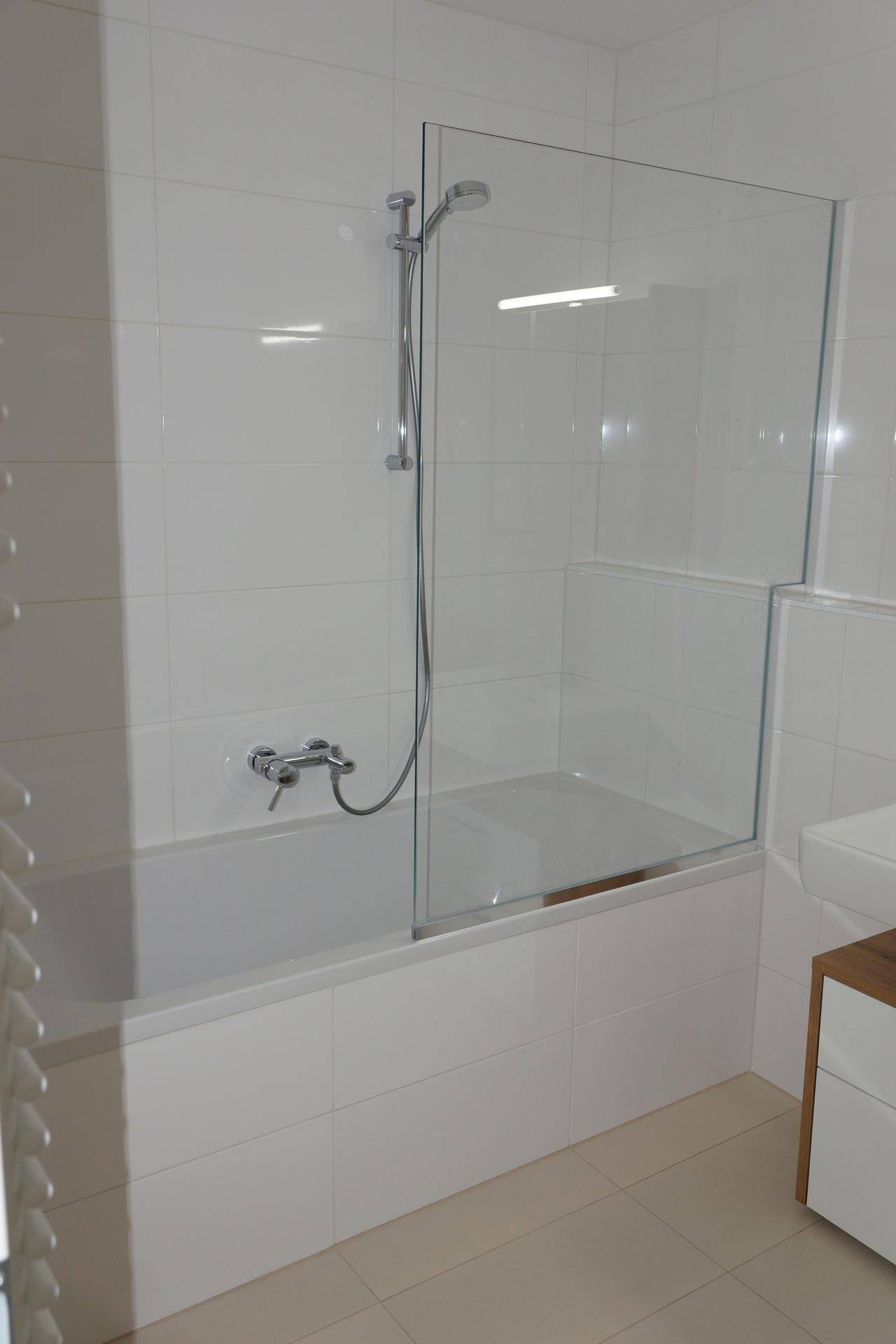 Modernes Badezimmer mit weißen Fliesen, einer Badewanne mit Glasabtrennung und Duschfunktion.