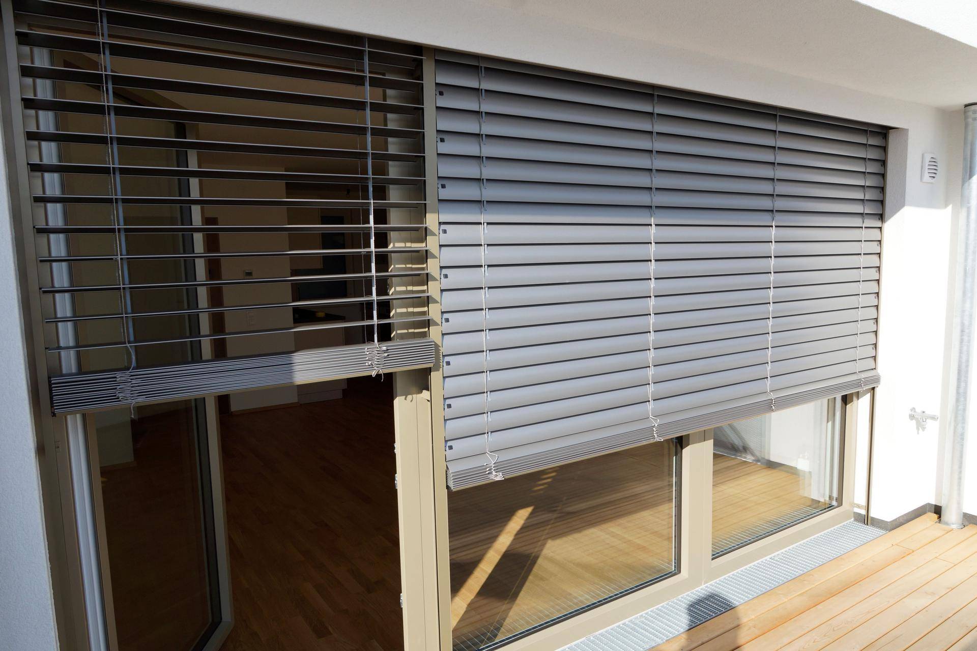 Große Fenster mit Außenjalousien bieten Zugang zu einem Balkon mit Holzboden und Blick in den Wohnbereich.