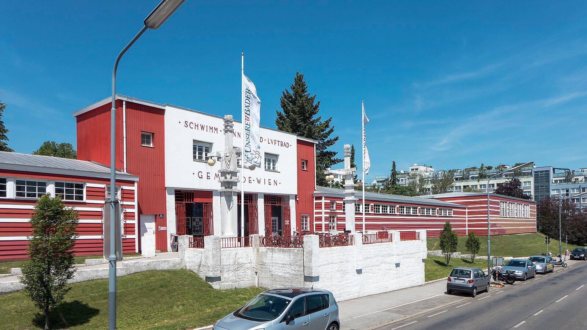 Außenansicht des Schwimmbads der Gemeinde Wien mit markanter rot-weißer Fassade unter blauem Himmel.