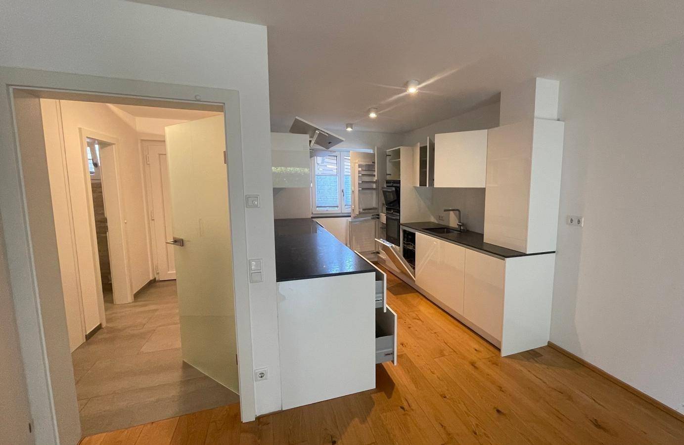 Moderne Einbauküche mit weißen Hochglanzfronten, dunkler Arbeitsplatte und Holzboden.