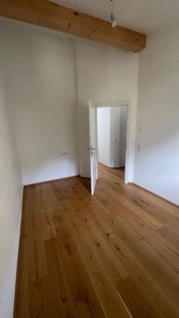 Leerer Raum mit Holzboden, weiß gestrichenen Wänden und sichtbarem Holzbalken.