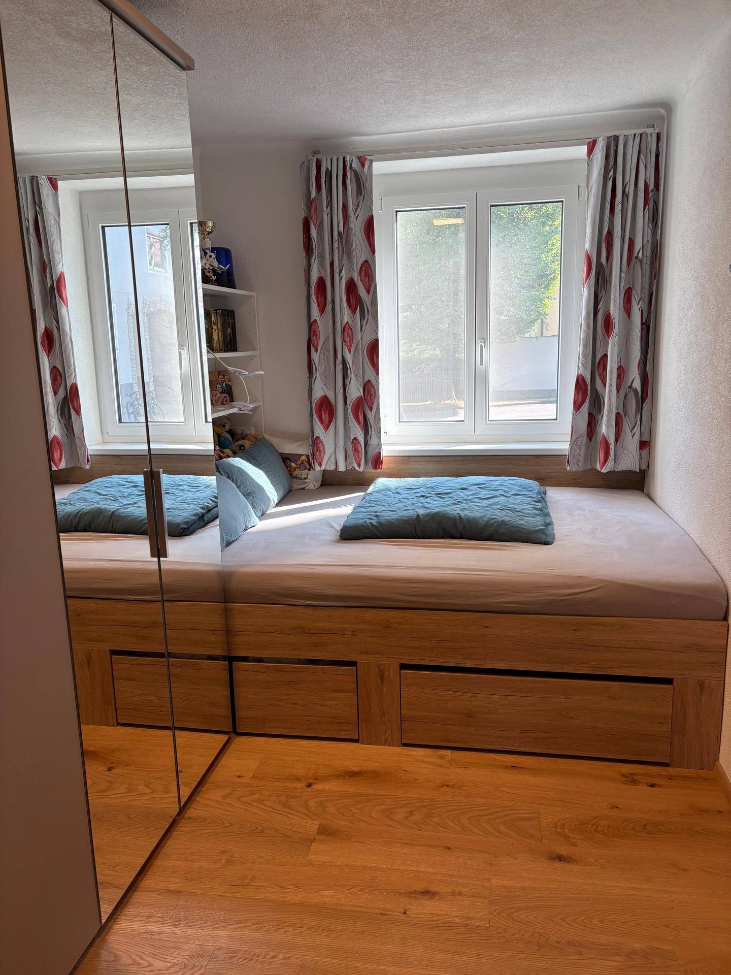 Gemütliches Schlafzimmer mit Holzbett, Parkettboden und großem Fenster, das viel Tageslicht hereinlässt.