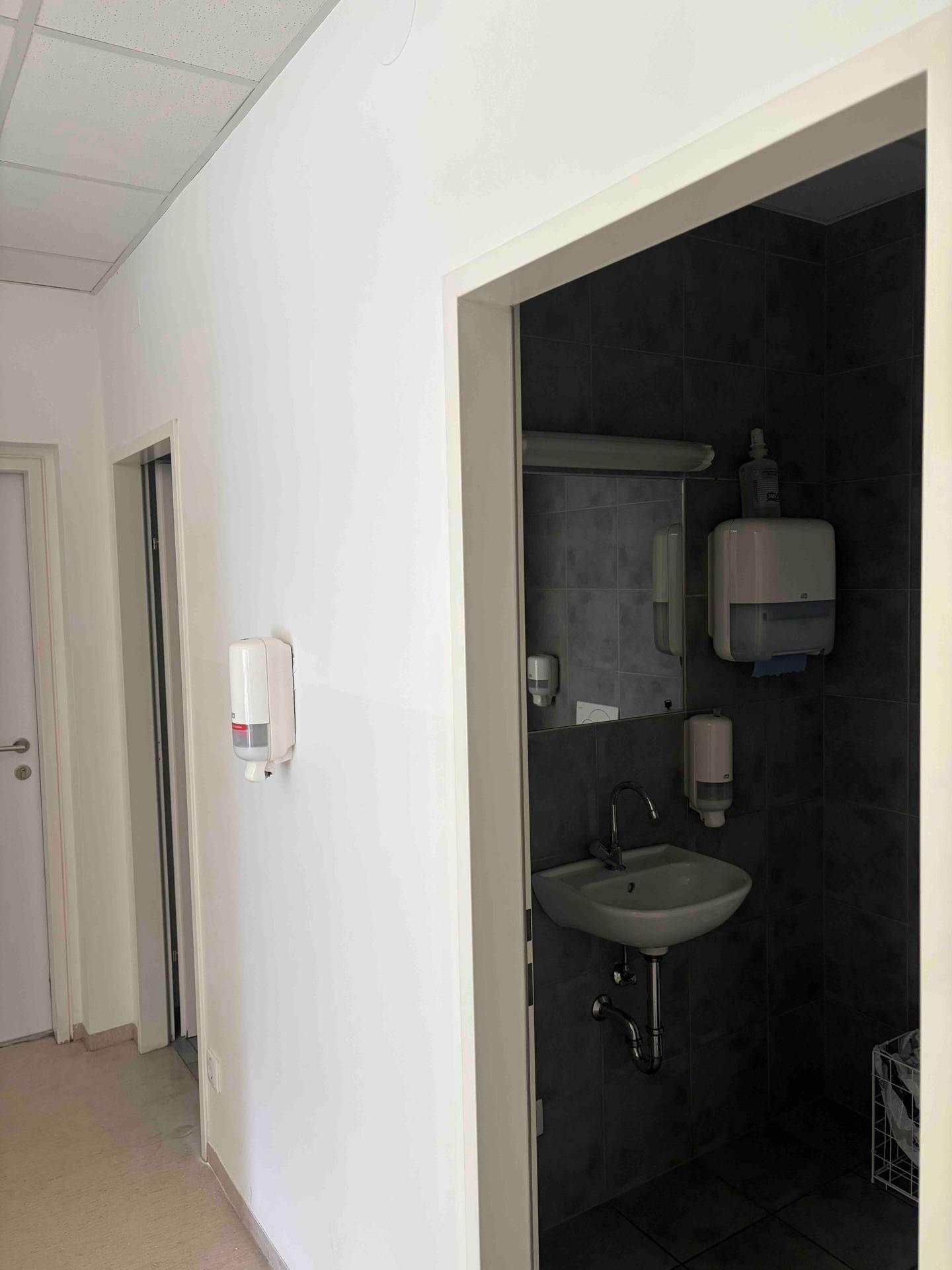 Kleiner Toilettenraum mit dunklen Fliesen, einem Waschbecken und Seifenspendern an der Wand.