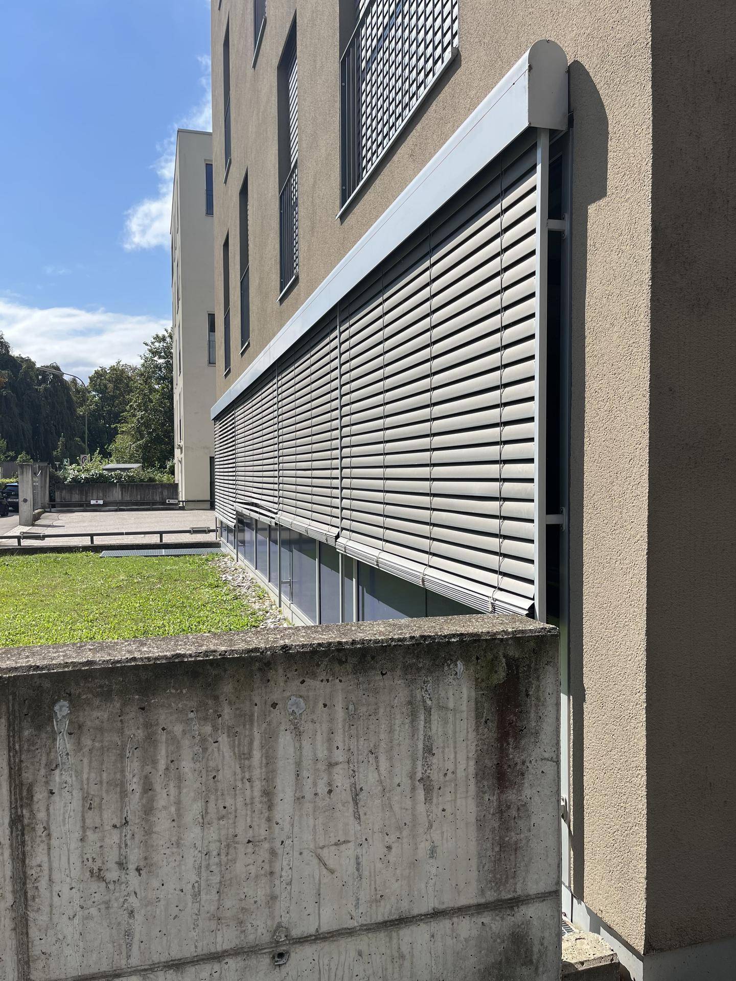 Nahaufnahme der Gebäudeseite mit geschlossenen Rollläden vor den Fenstern und einer Betonmauer im Vordergrund.