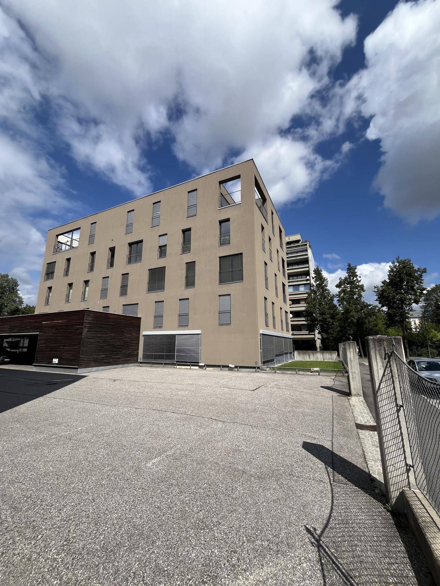 Modernes Gebäude mit mehreren Etagen und einer Parkfläche im Vordergrund unter bewölktem Himmel.