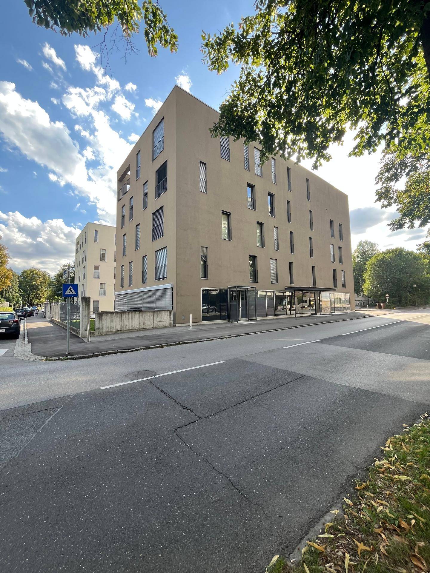 Modernes Eckgebäude mit mehreren Etagen und vielen Fenstern, umgeben von Bäumen und einer Straße.