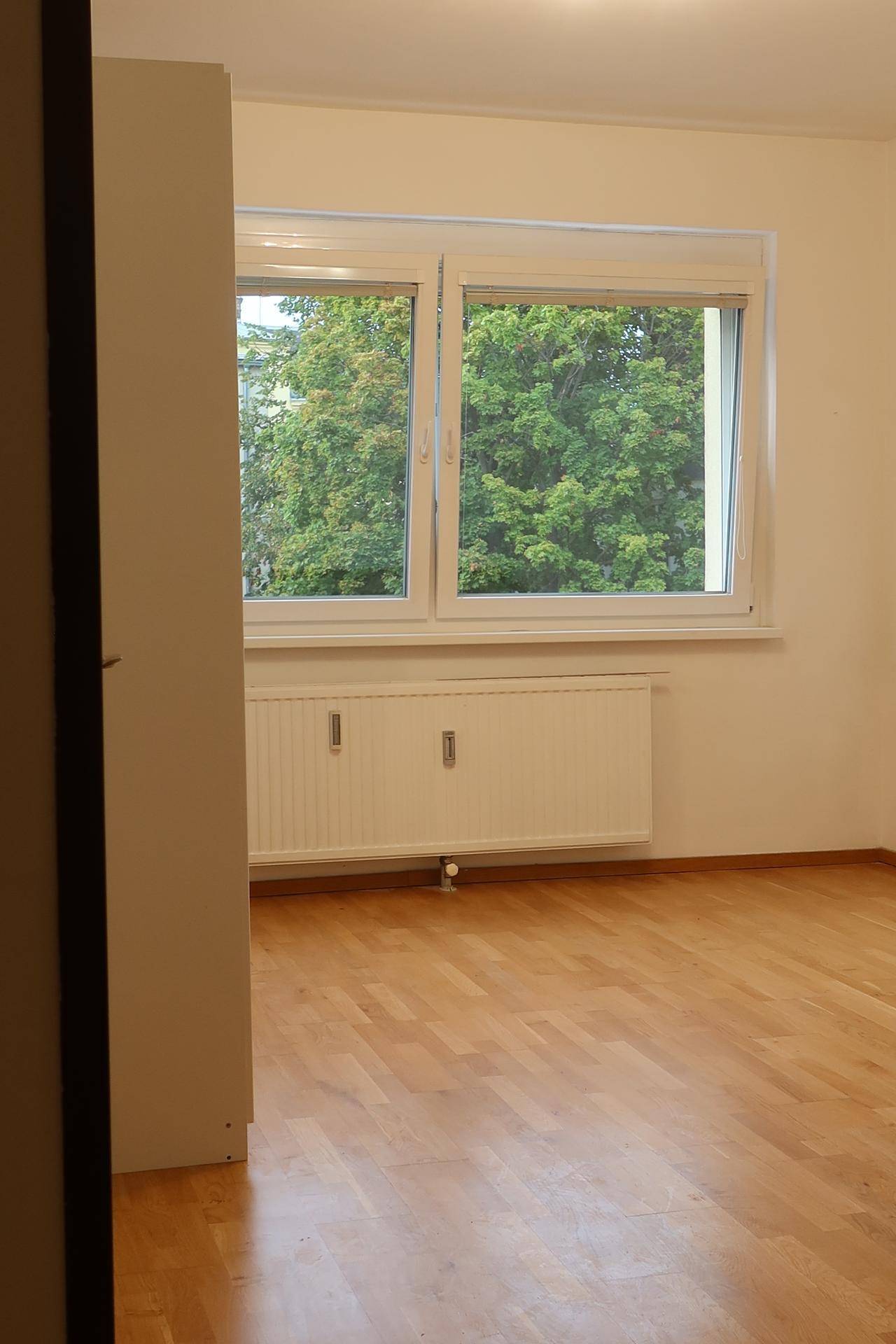 Geräumiges Zimmer mit Parkettboden und weitem Blick auf grüne Bäume durch das große Fenster.