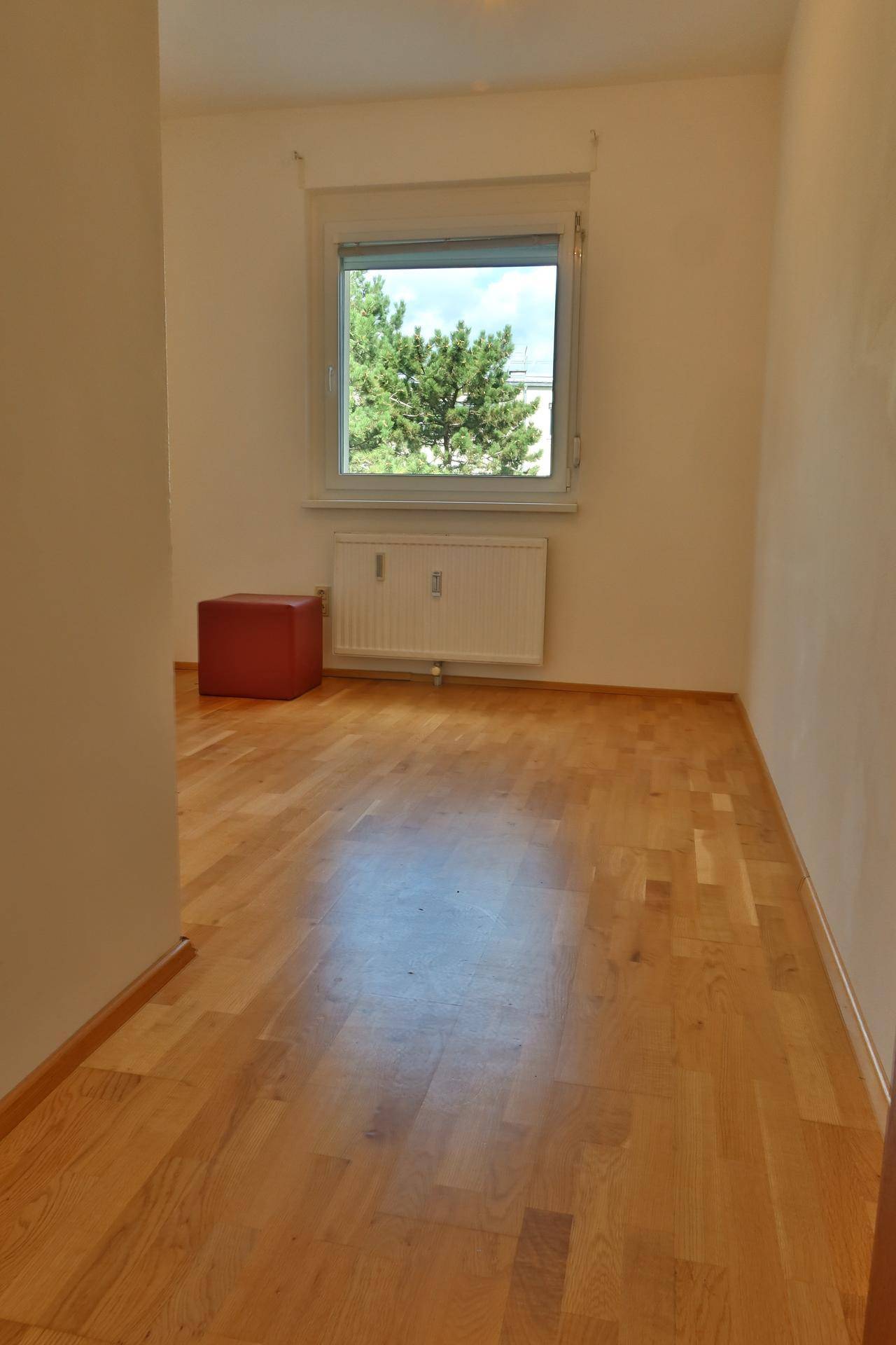 Leeres Zimmer mit hellem Holzboden, großem Fenster und Blick ins Grüne.