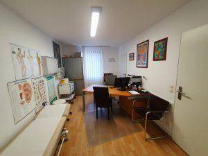 Geräumiges Untersuchungszimmer mit Holzboden, Schreibtisch und medizinischen Wandtafeln.