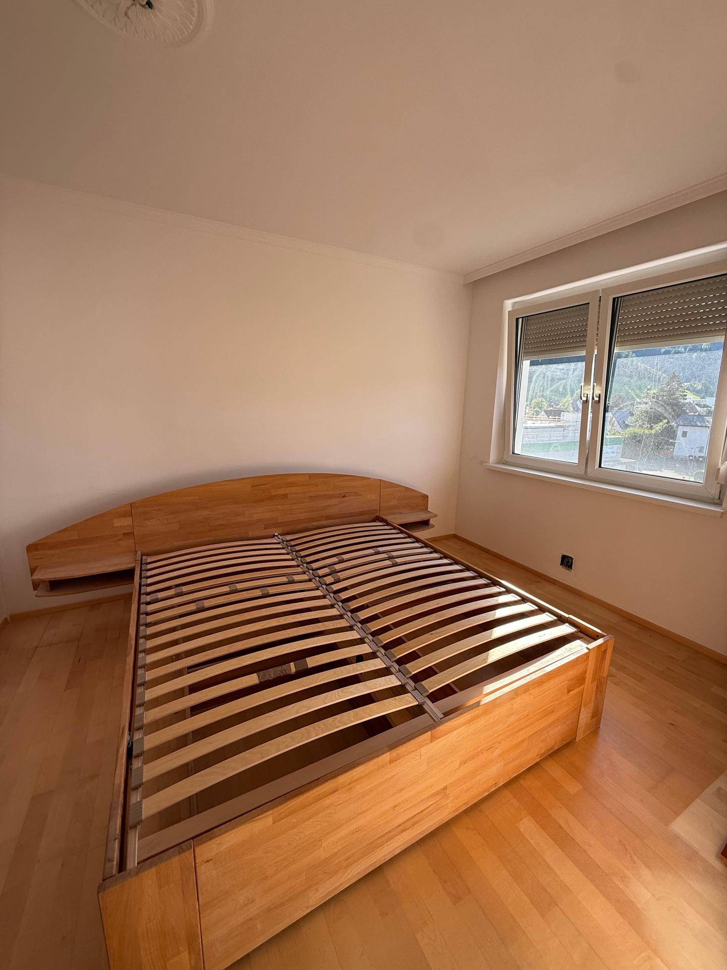 Schlafzimmer mit hellem Holzfußboden, großem Fenster und einem stabilen Bettrahmen.