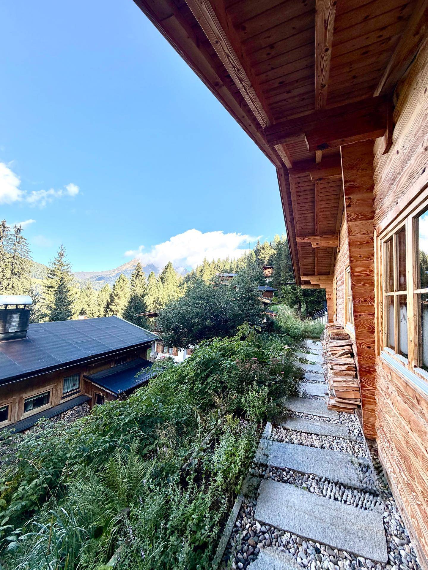 Steinplattenweg entlang der Holzfassade eines Chalets mit Blick auf die grüne Umgebung.