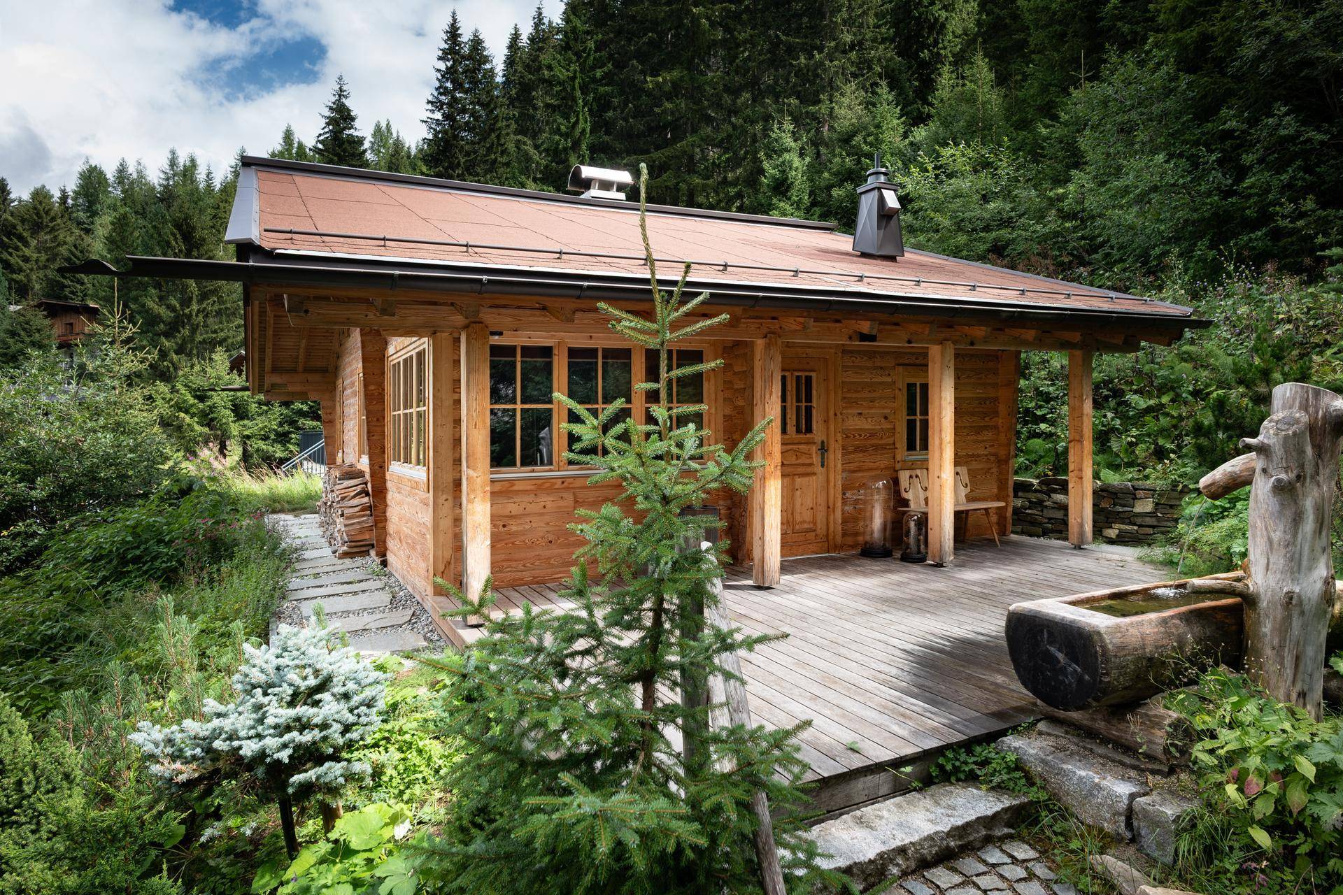 Charmantes Holzhaus mit Terrasse und rustikalem Wasserbrunnen, umgeben von grüner Natur.