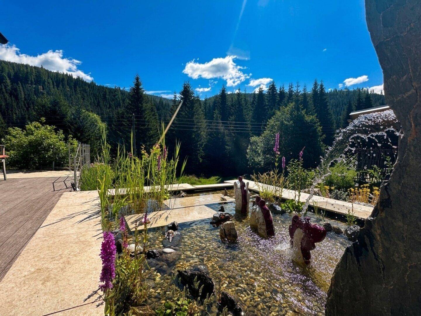 Dekorativer Wasserlauf im Garten mit Steinen und Pflanzen, umgeben von üppiger Natur und Wald.