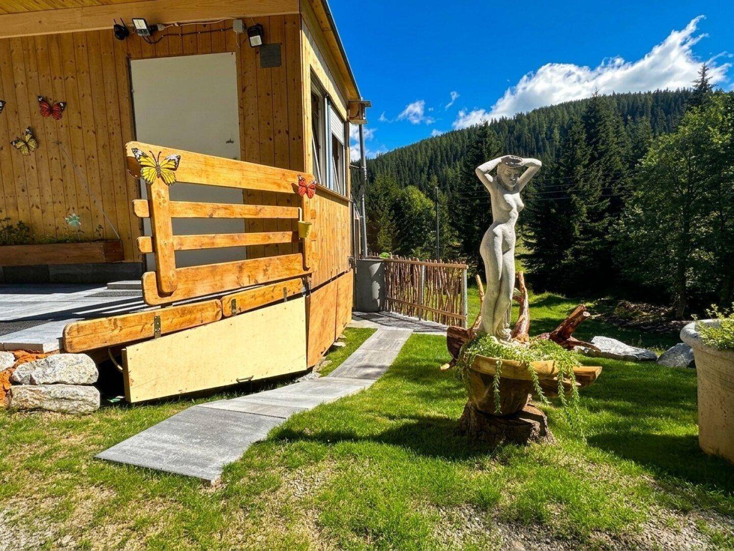 Rustikales Holzhaus mit kleiner Terrasse und dekorativer Statue im Vorgarten, umgeben von Wald.