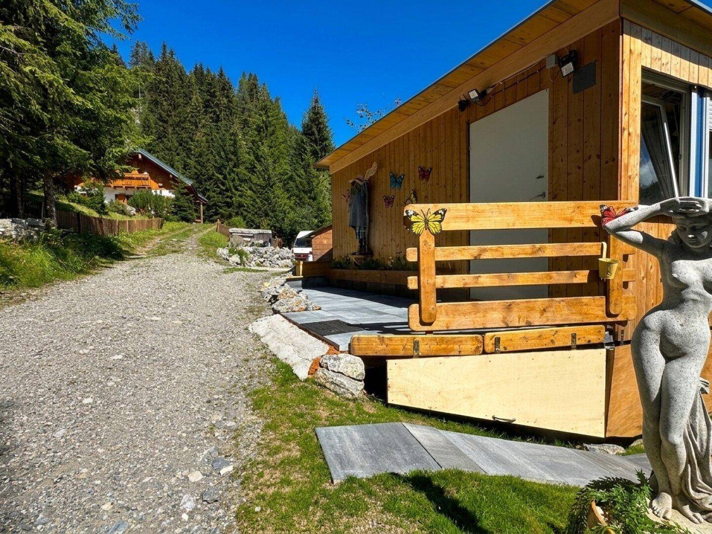 Holzhaus mit Schotterweg und dekorativer Statue, eingebettet in eine bewaldete Berglandschaft.