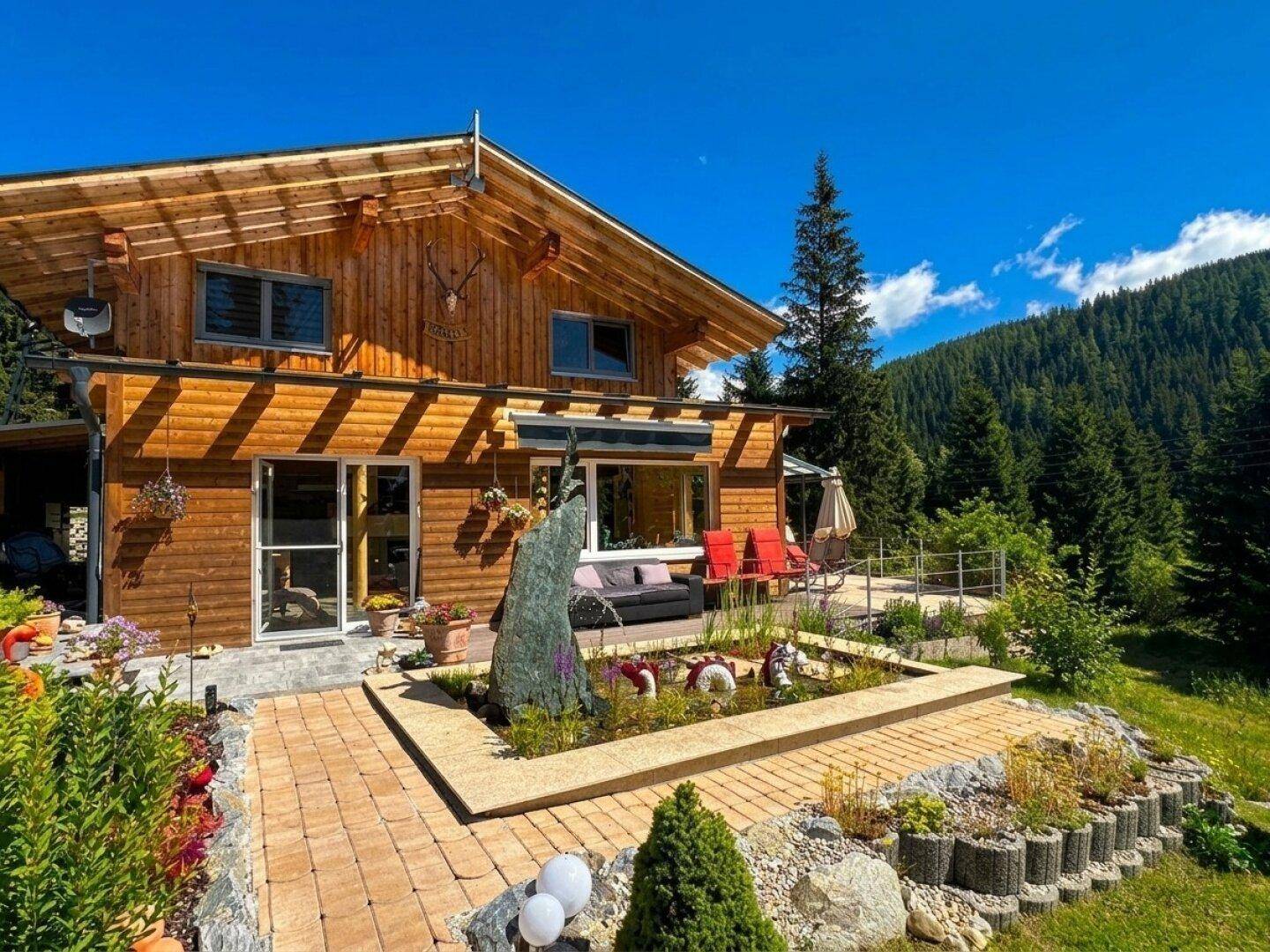 Einladende Terrasse vor dem Holzhaus mit Wasserspiel, Gartenbeeten und Bergblick.