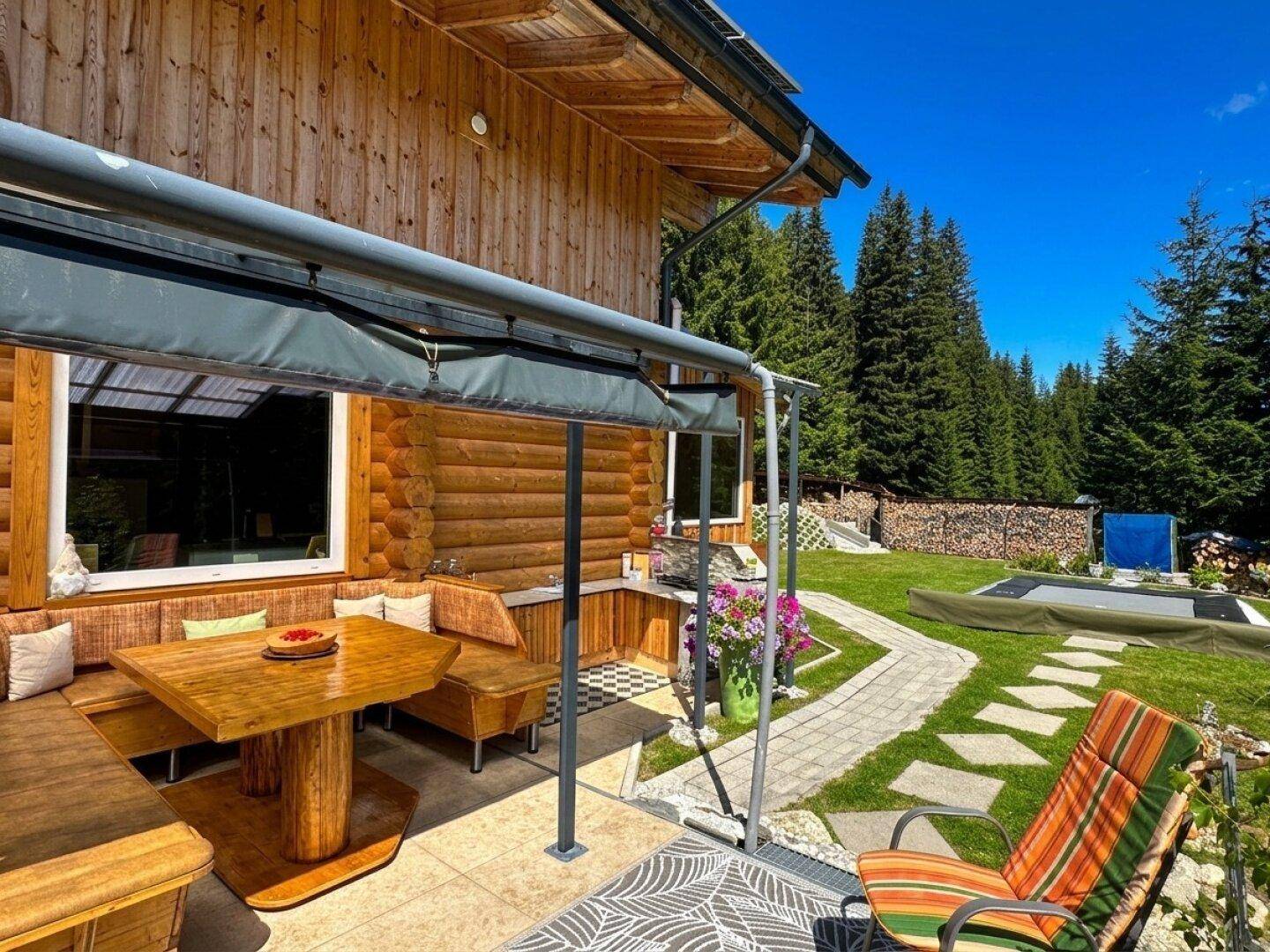 Überdachter Essbereich im Freien mit Holztisch und Bänken, angrenzend an den Garten mit Trampolin.