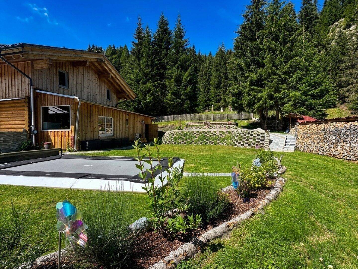 Blühendes Blumenbeet im Vordergrund eines gepflegten Rasens mit dem Holzhaus im Hintergrund.