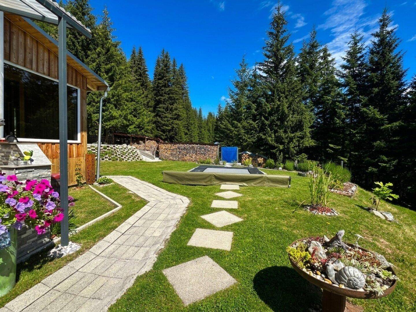 Gepflegter Garten mit gepflastertem Weg, Trittsteinen und Trampolin, umgeben von Wald.
