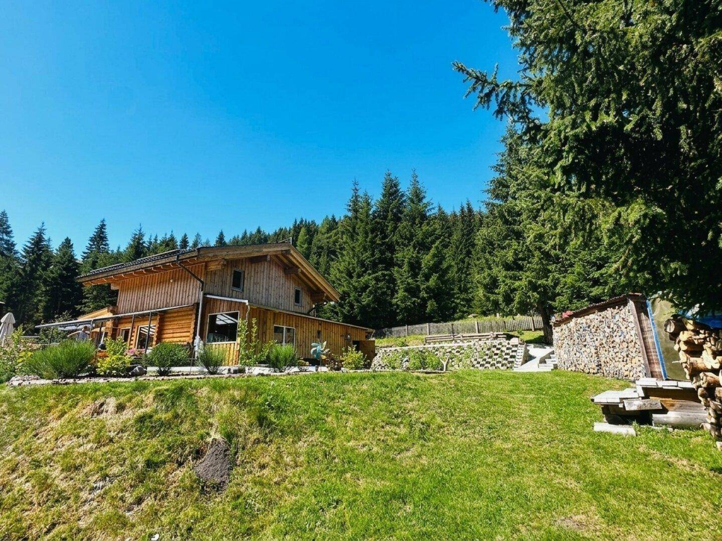 Rustikales Holzhaus auf einem Hügel mit weitem Rasen und Holzstapel, umgeben von Wald.